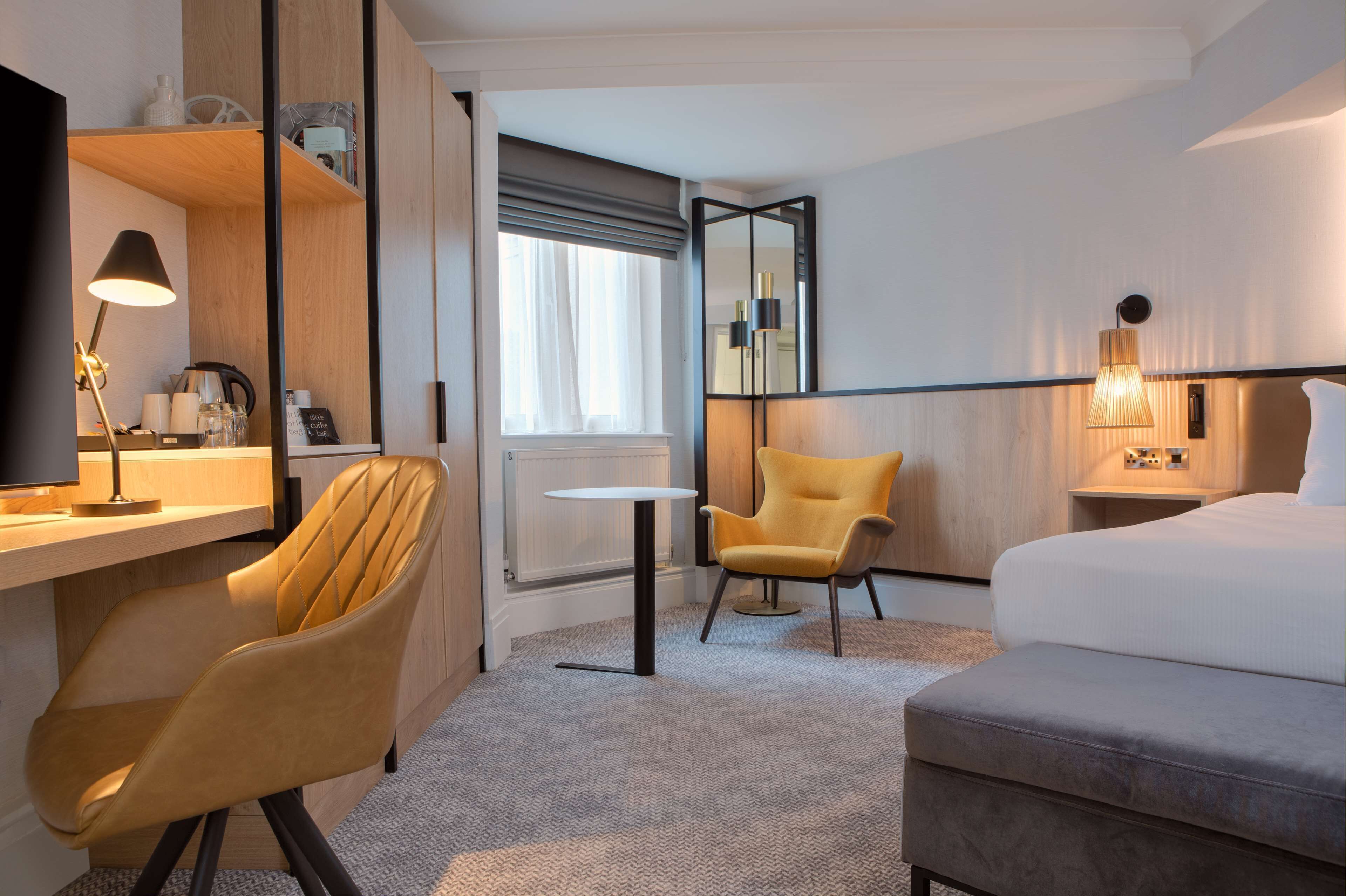 A hotel room with a bed, a desk with a lamp, a chair, a small table, and a large mirror.