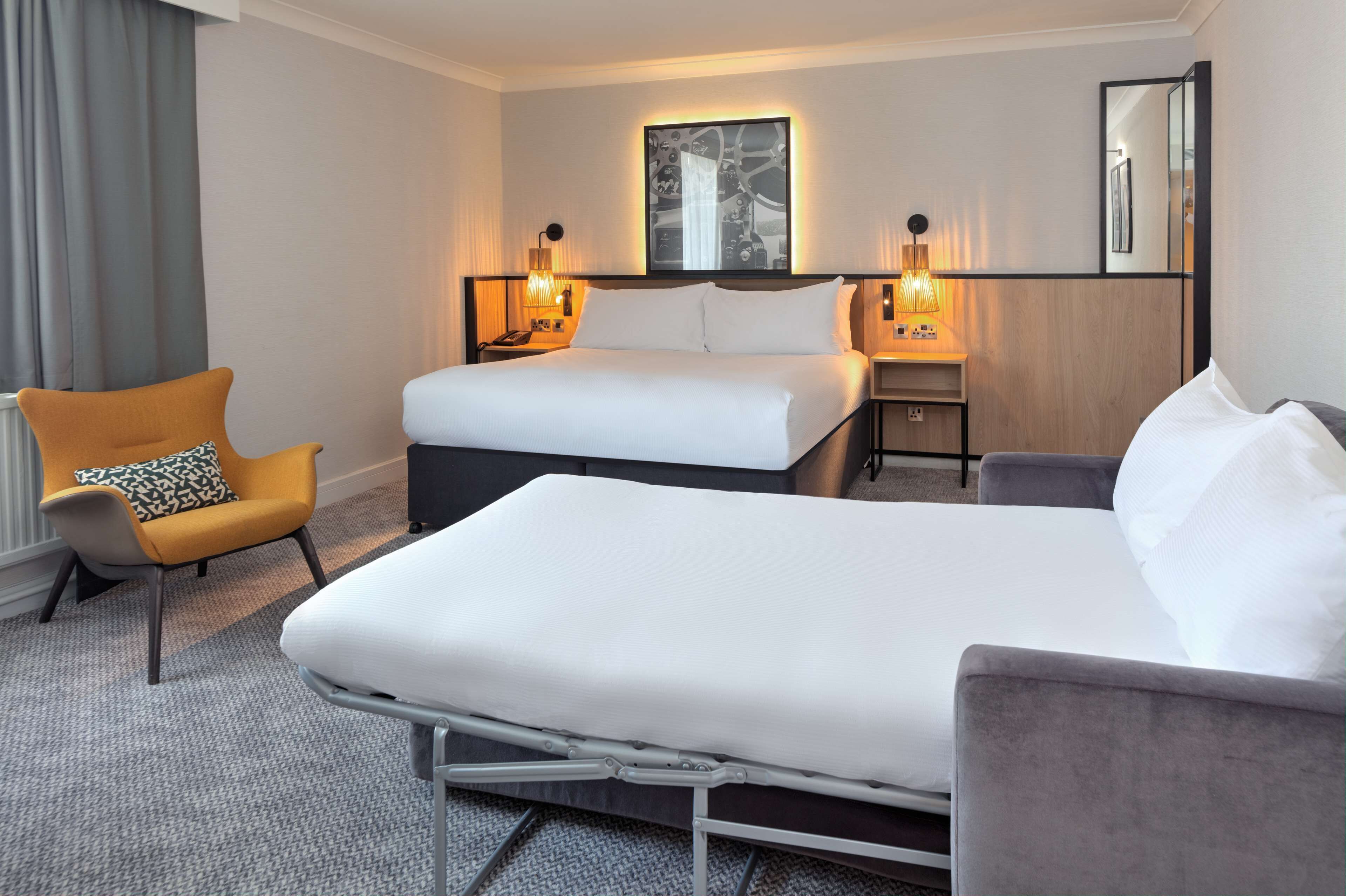 Habitación de hotel con una cama grande, una cama más pequeña, una silla y una mesita de noche con lámparas.