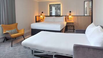 Habitación de hotel con una cama grande, una cama más pequeña, una silla y una mesita de noche con lámparas.