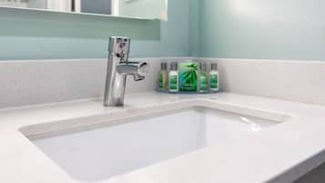 Uma pia de banheiro com uma torneira cromada e um espelho acima. Há várias garrafas de shampoo verdes na bancada.