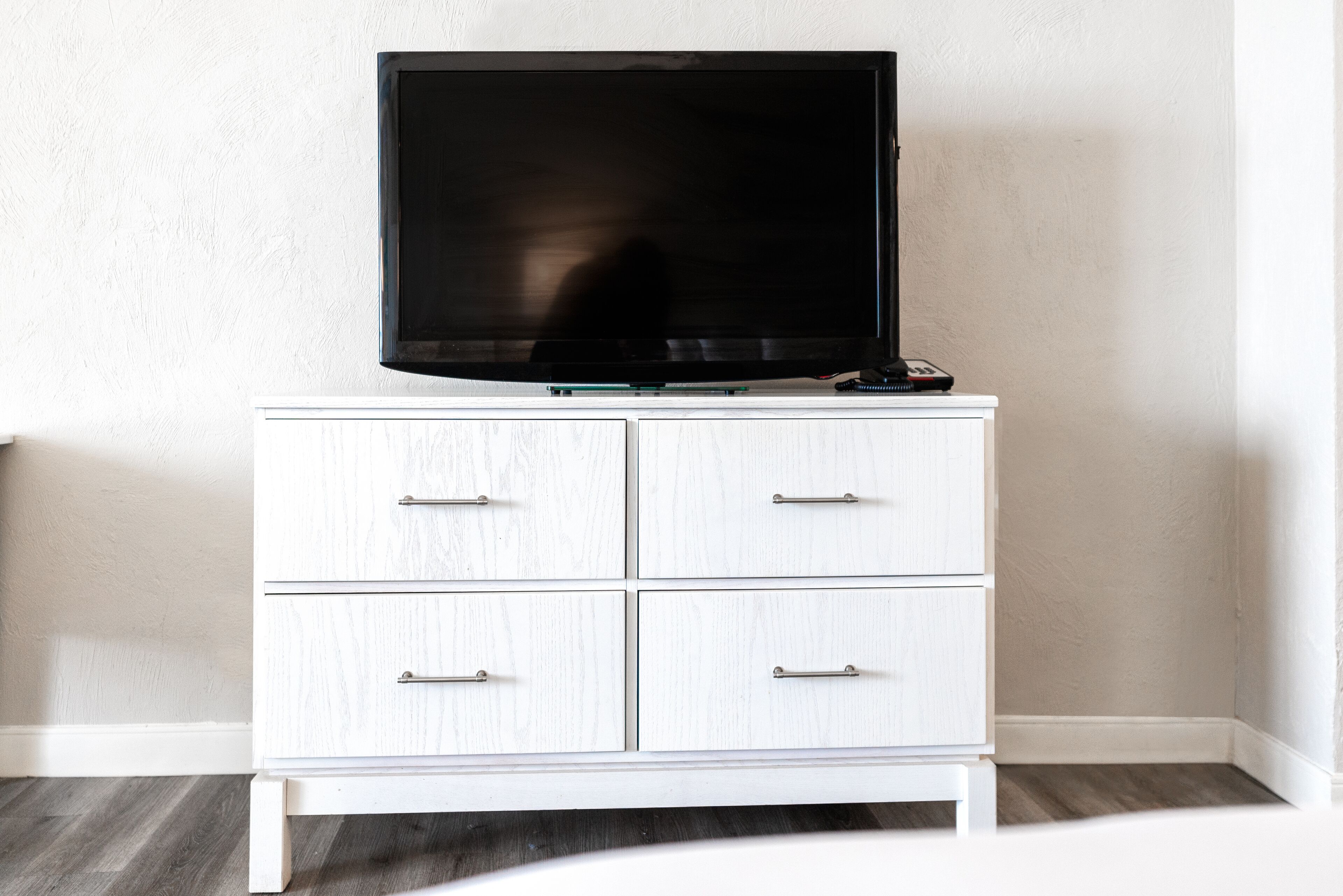 A white dresser with a flat-screen TV on top, drawers with metal handles, and a wooden floor.