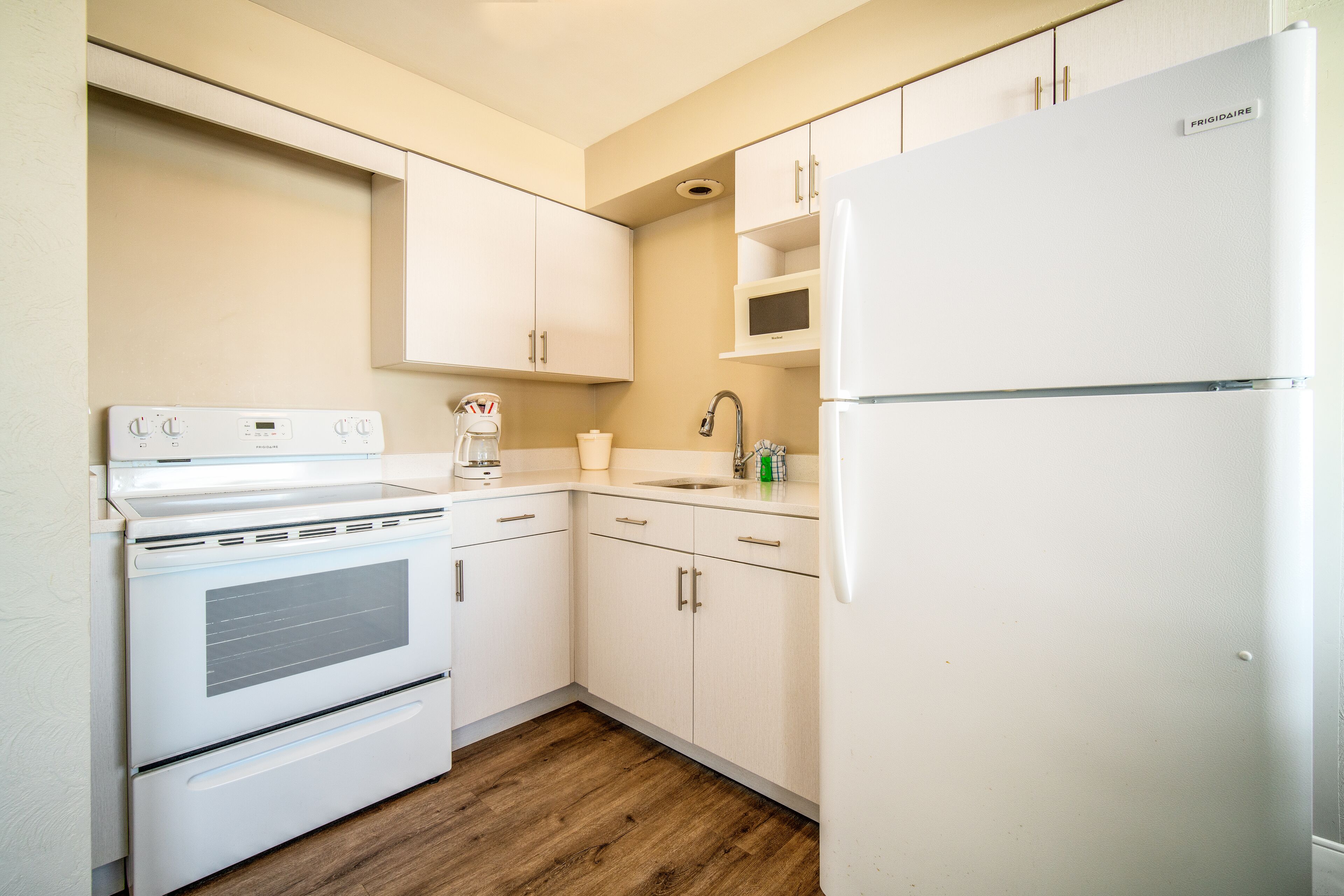 A kitchen with white appliances, wooden flooring, and light-colored cabinets.