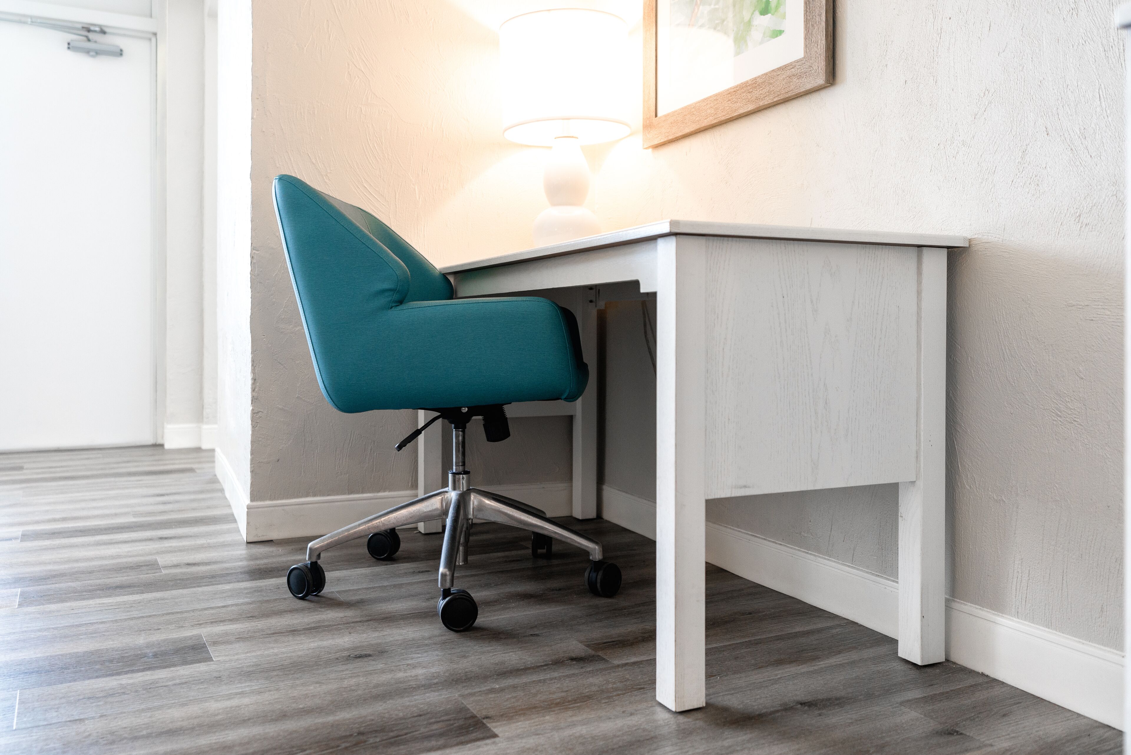 Une chaise de bureau turquoise à roulettes, un bureau blanc et une lampe posée sur le bureau.