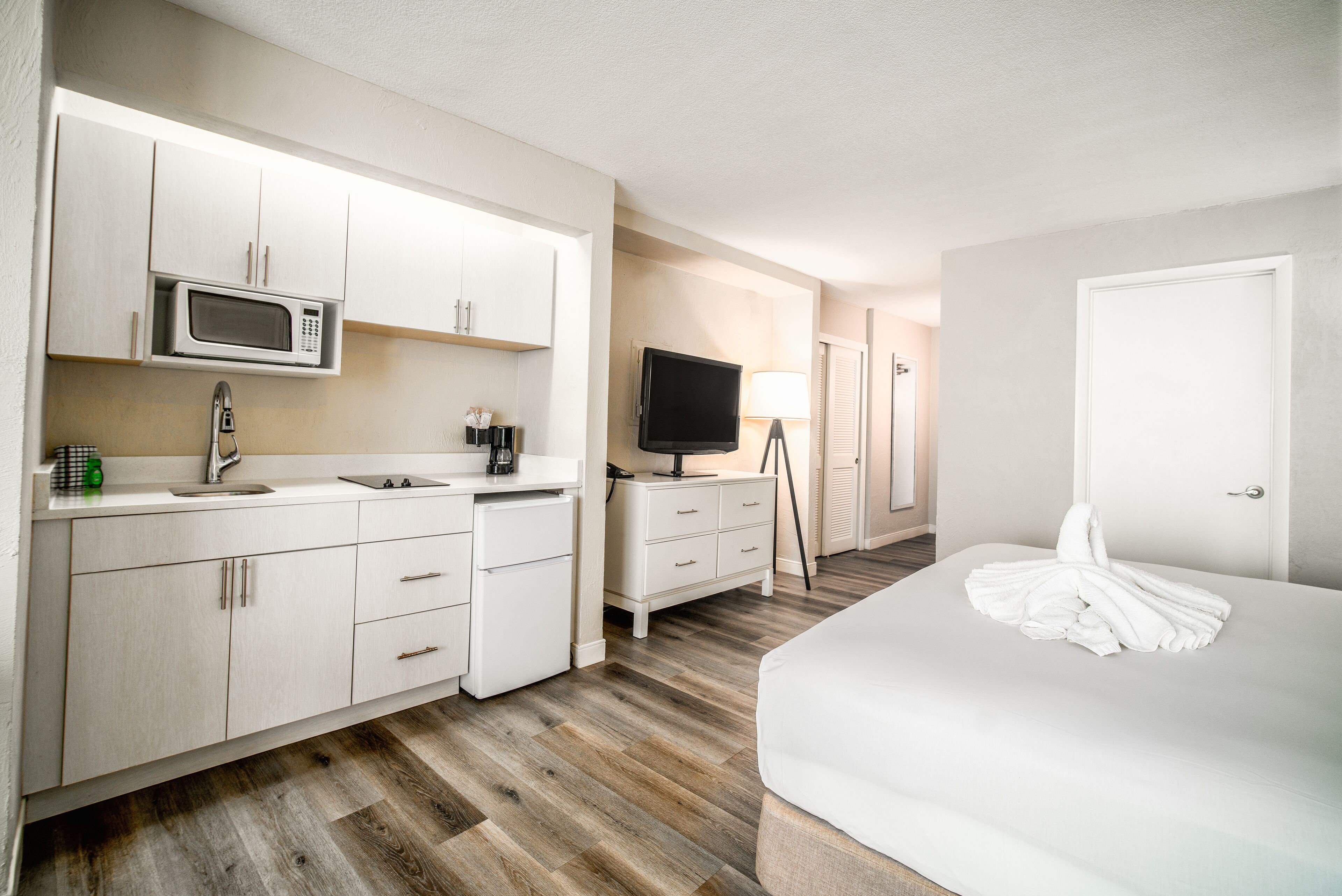 A hotel room with a kitchenette, a bed, a TV, and a lamp.