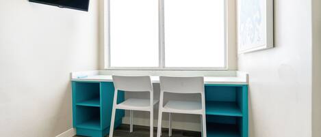 A room with a white desk, two white chairs, teal shelving, a flat-screen TV, and a large window.