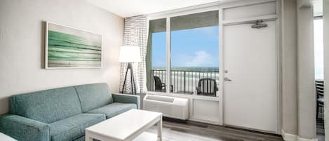 A living room with a teal sofa, a white coffee table, a large window with a view of the ocean, and a sliding glass door leading to a balcony.