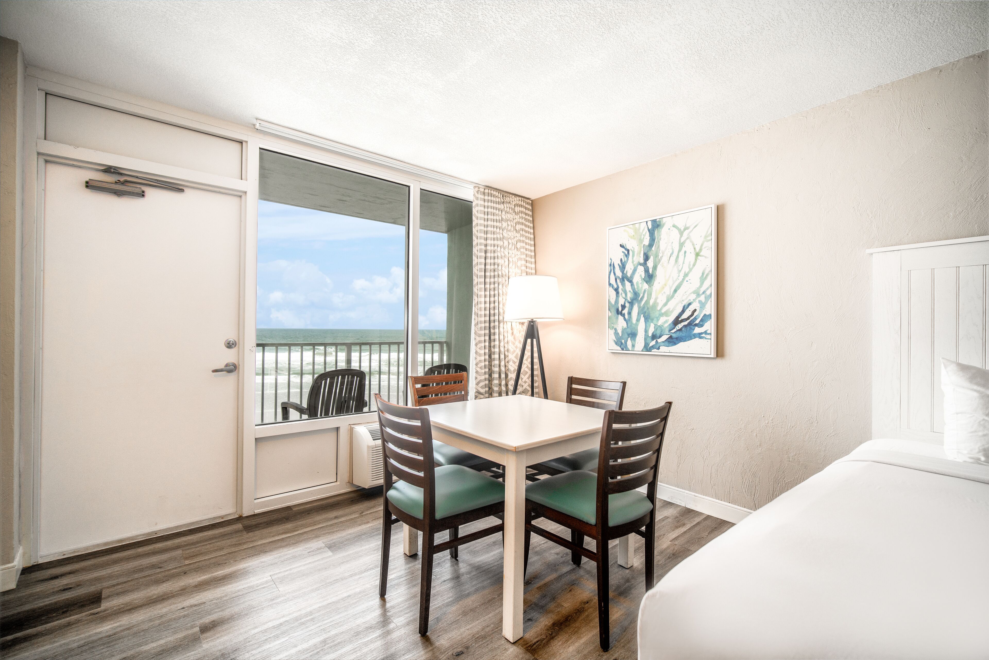 A hotel room with a balcony, a dining table with chairs, a bed, and a painting on the wall.