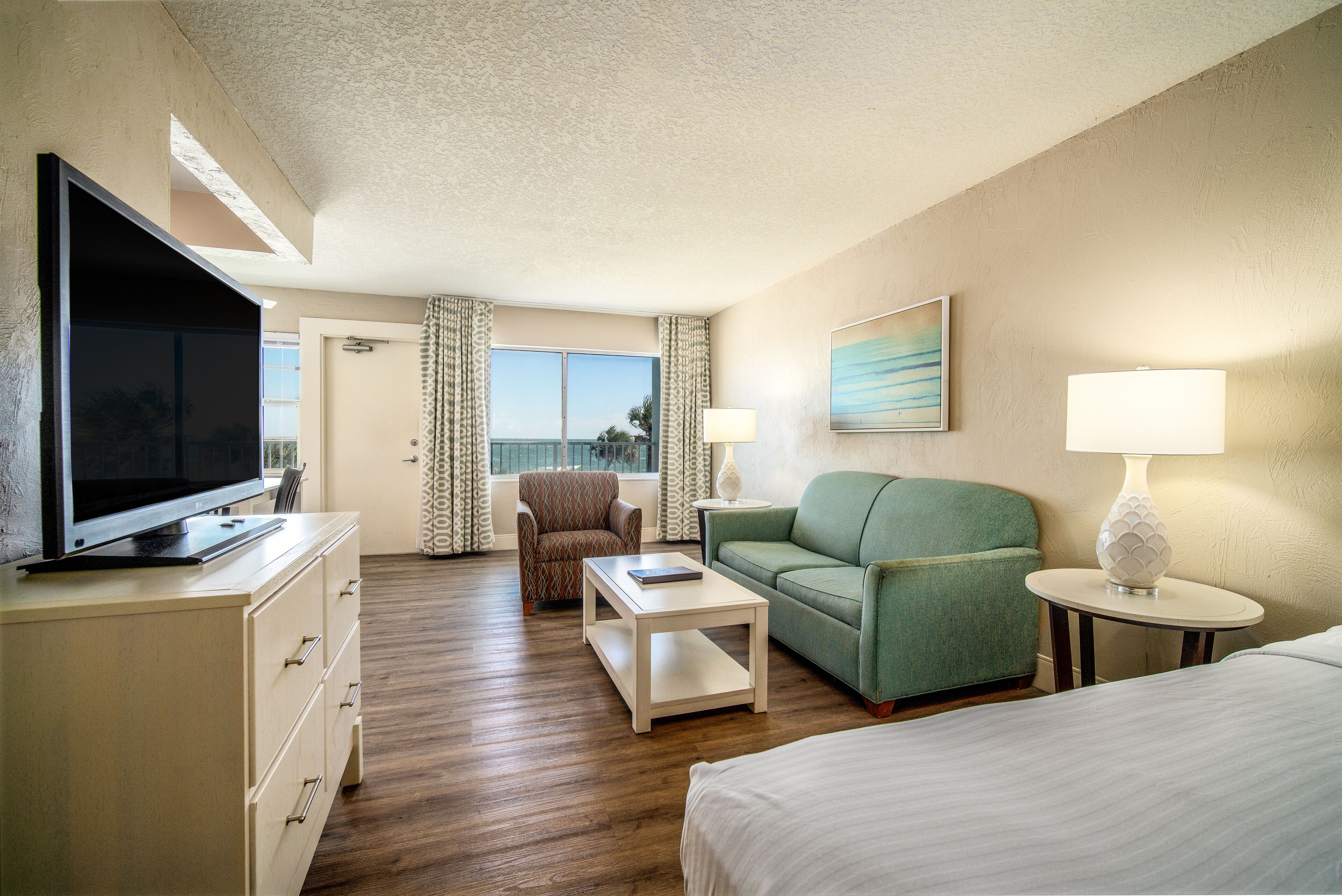 Une chambre d’hôtel avec une télévision posée sur une commode, un canapé vert, une table basse blanche, une lampe sur une table d’appoint et un lit.