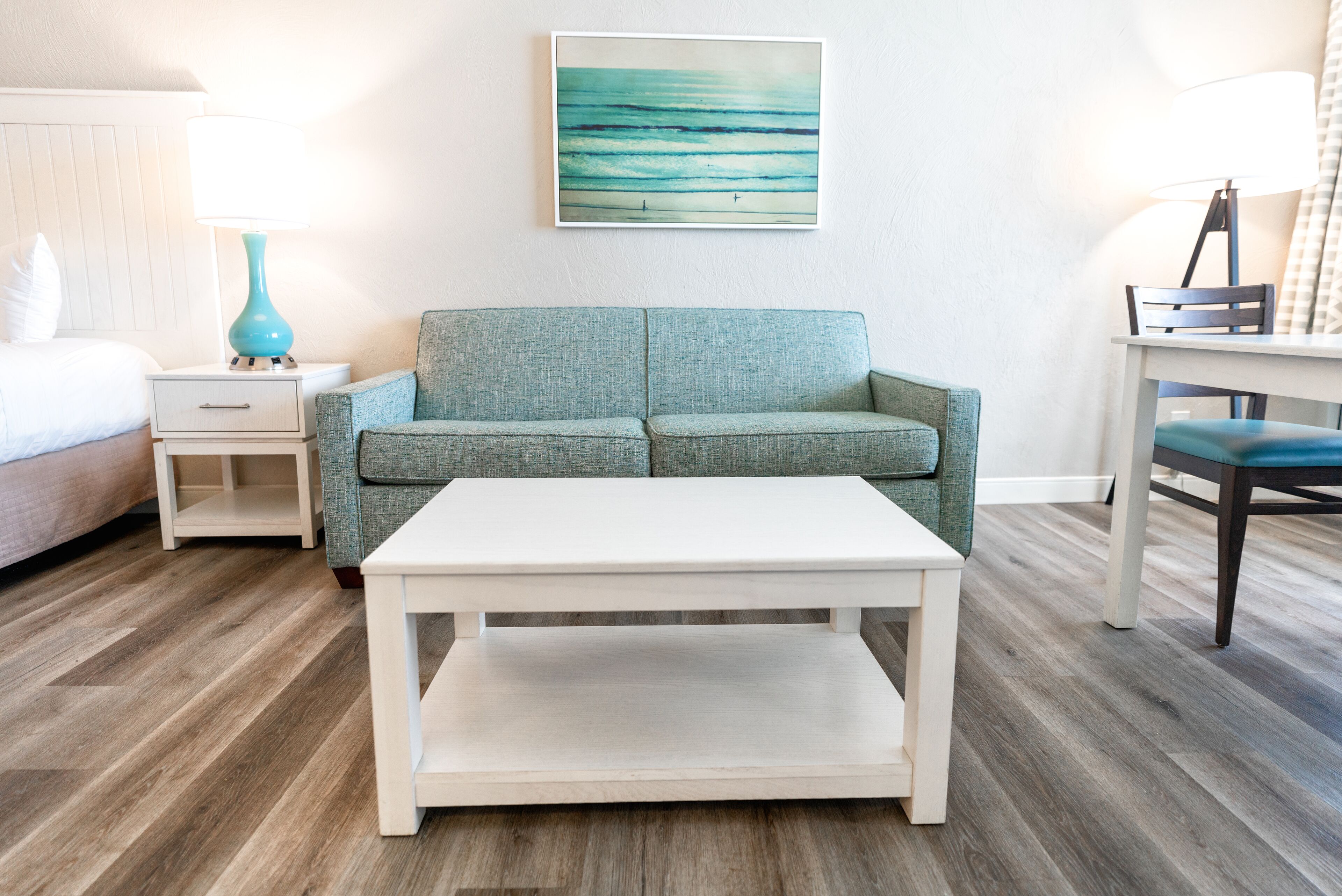 Quarto com sofá verde-água, mesa de centro branca, cama com travesseiro branco, mesa de cabeceira com abajur verde-água e um quadro na parede.