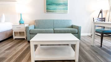 A room with a teal sofa, a white coffee table, a bed with a white pillow, a nightstand with a teal lamp, and a painting on the wall.
