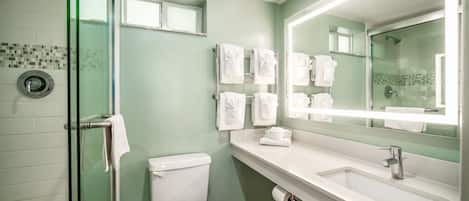 Baño con cabina de ducha de vidrio, inodoro, lavabo con espejo y varios toallas en un perchero.