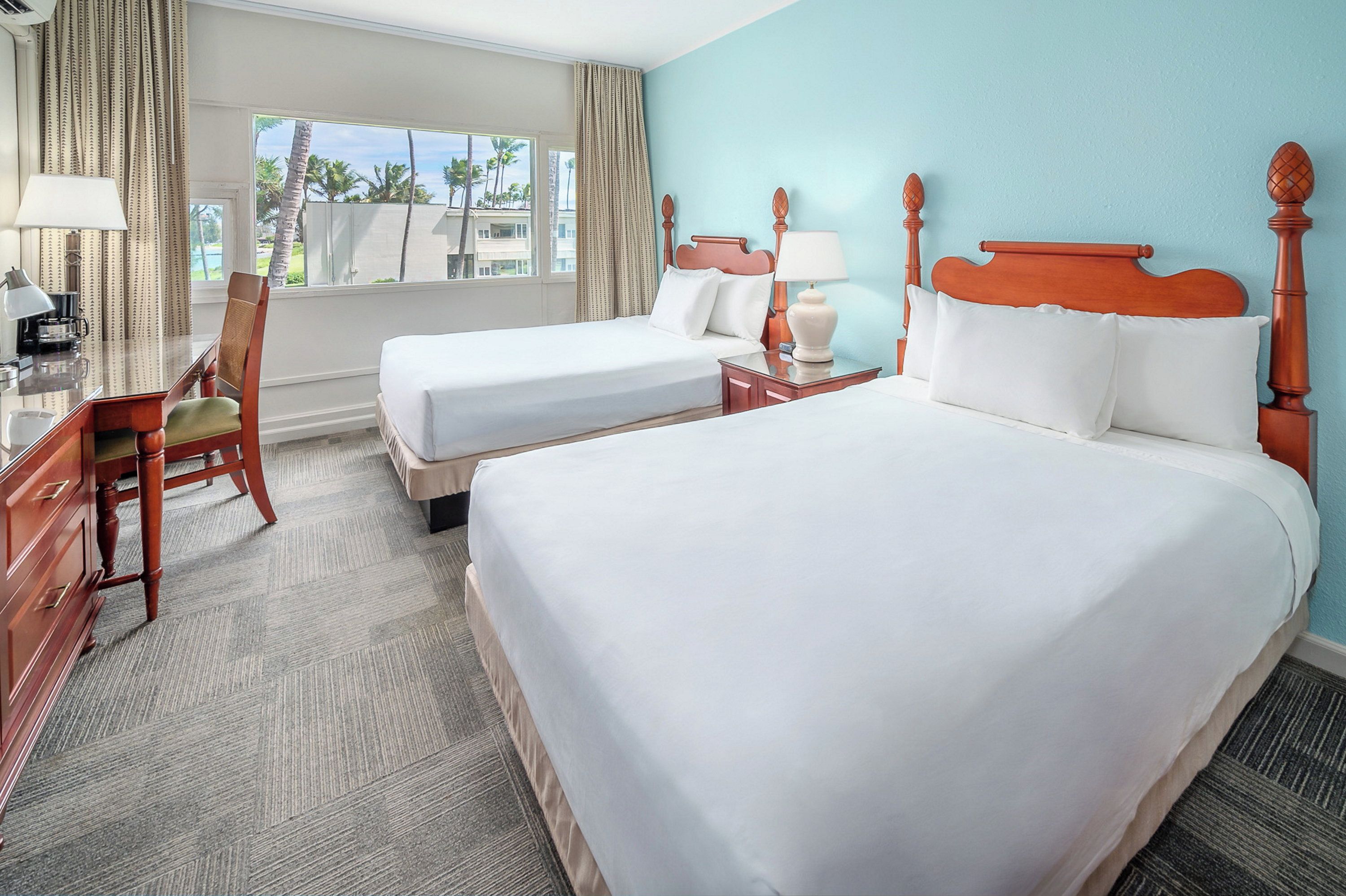 A hotel room with two beds, a desk, a chair, and a view of palm trees.