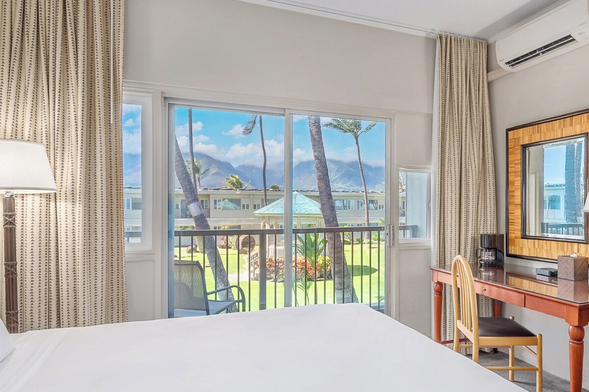 Une chambre d’hôtel avec un grand lit, un bureau, une chaise et un balcon offrant une vue sur les palmiers et les montagnes.