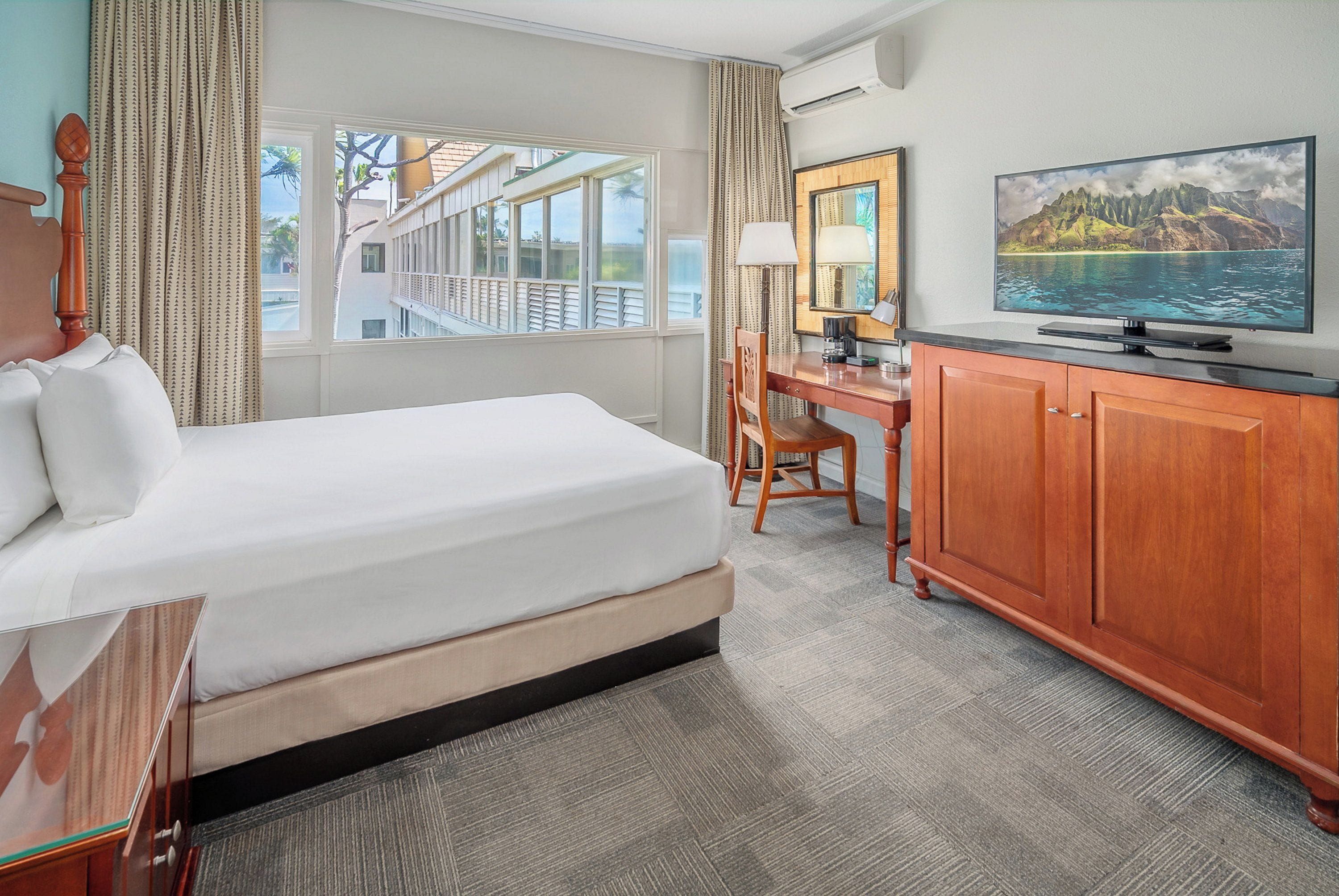 Habitación de hotel con cama, escritorio, silla, televisión y un ventanal con vistas a un balcón.