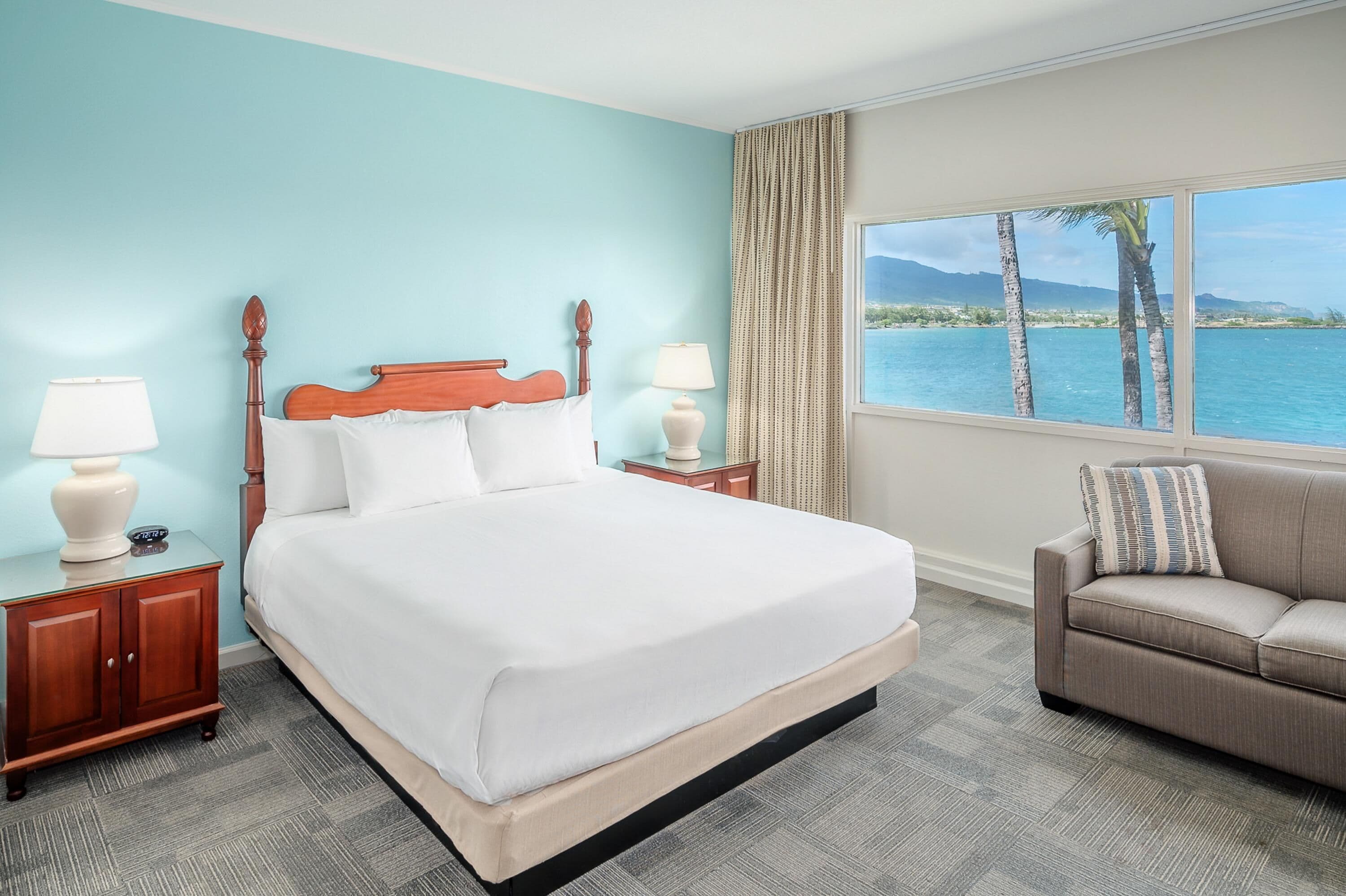 Habitación de hotel con una cama grande, dos mesitas de noche, un sofá, un ventanal con vista al mar y palmeras, y una pared de color azul claro.