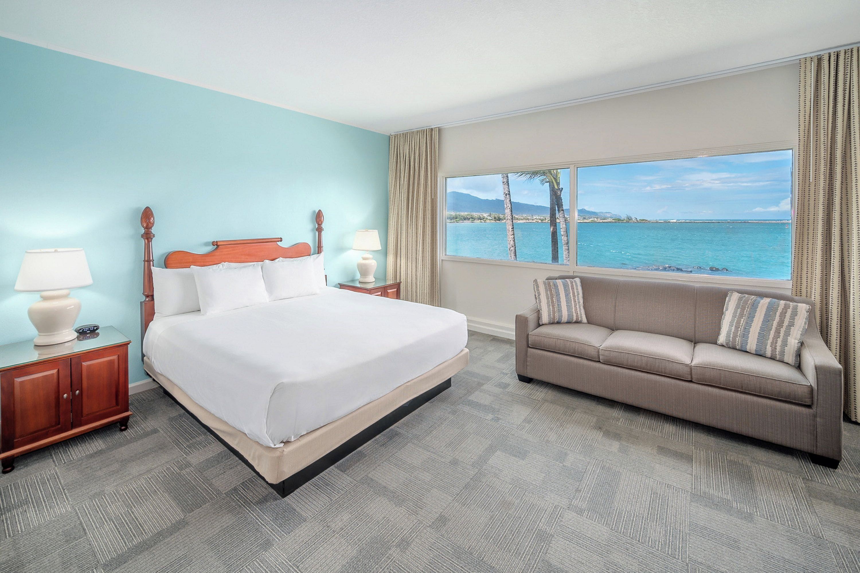 Une chambre d’hôtel avec un lit, un canapé, une table de chevet, une lampe et une fenêtre donnant sur la mer.