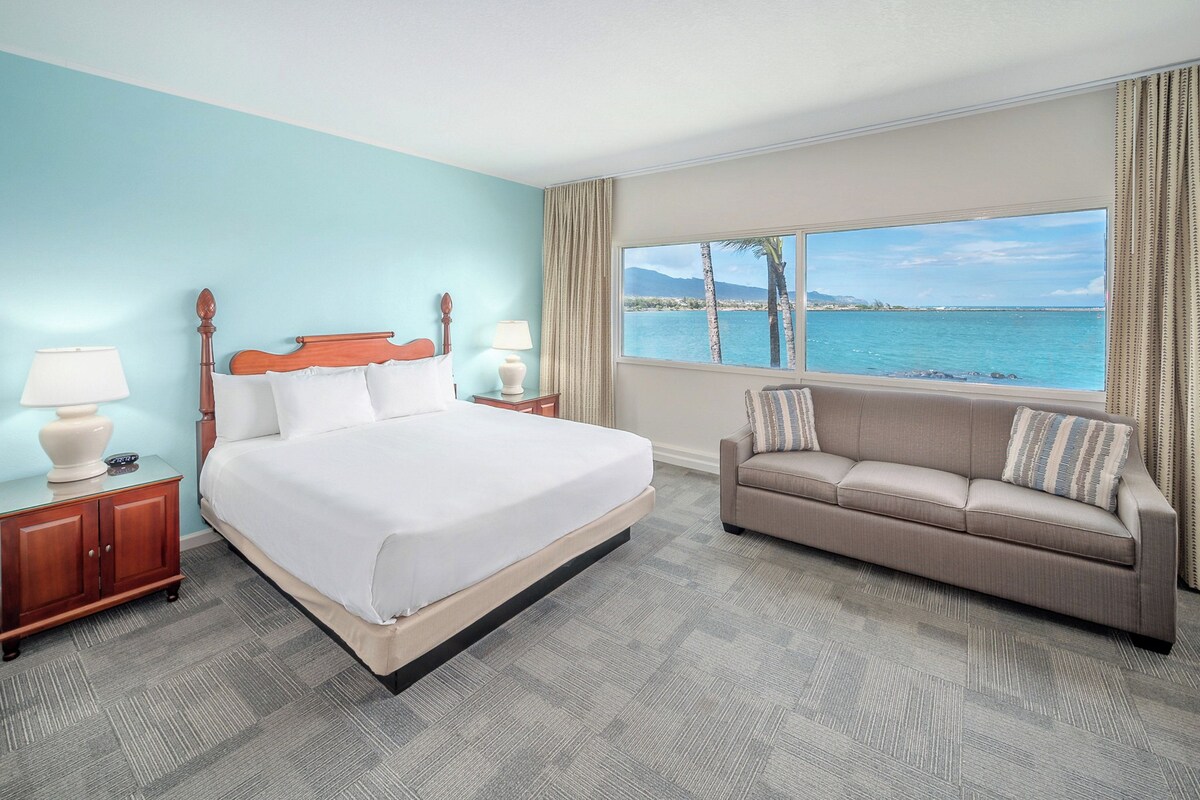 A hotel room with a bed, a sofa, a nightstand, a lamp, and a window with a view of the sea.