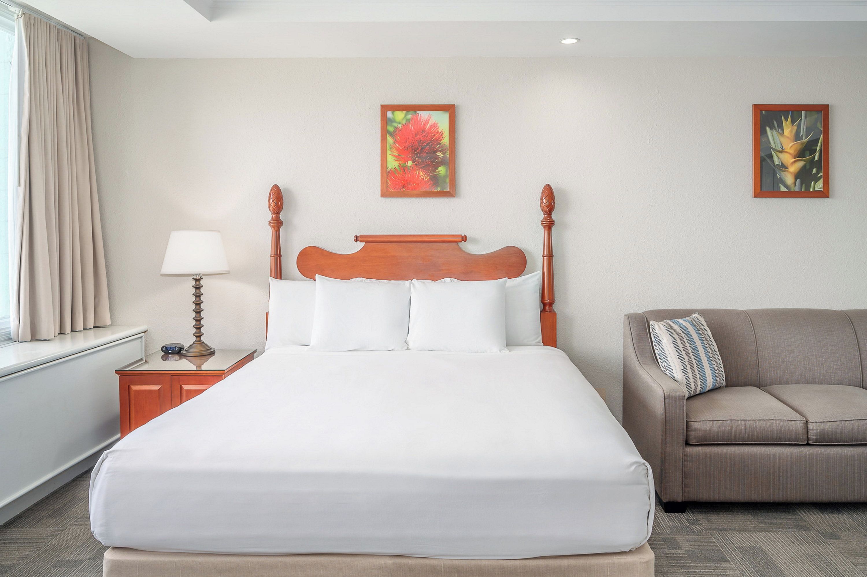 Una habitación de hotel con una cama grande, una mesita de noche con lámpara, un sofá y dos cuadros colgados en la pared.