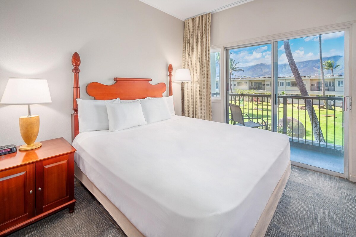 Un cuarto de hotel con una cama grande, una mesita de noche de madera con una lámpara y vista a un paisaje tropical a través de la ventana.