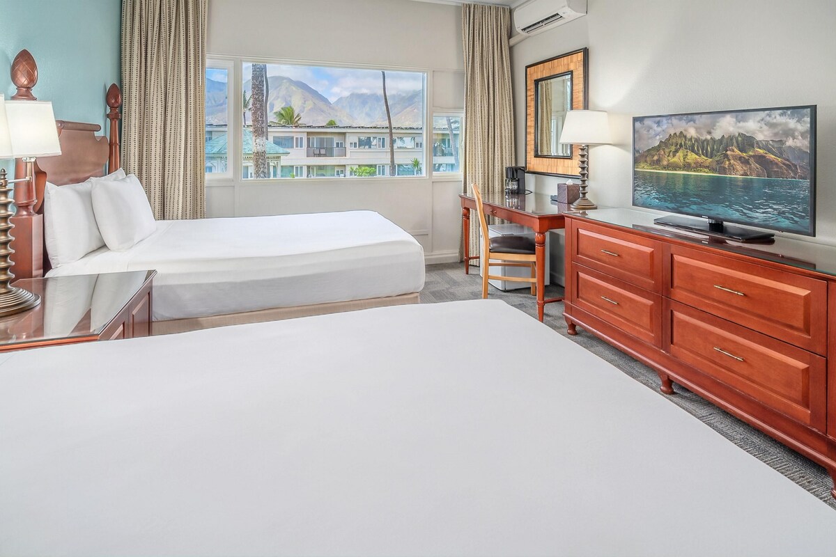 A hotel room with a bed, a desk with a chair, a television, and a view of mountains and a lake.