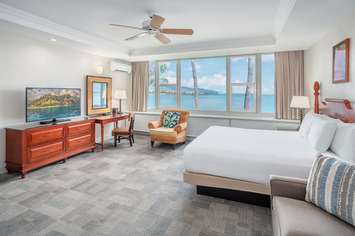 A hotel room with a bed, a TV, a desk, a chair, and a large window with a view of the sea.