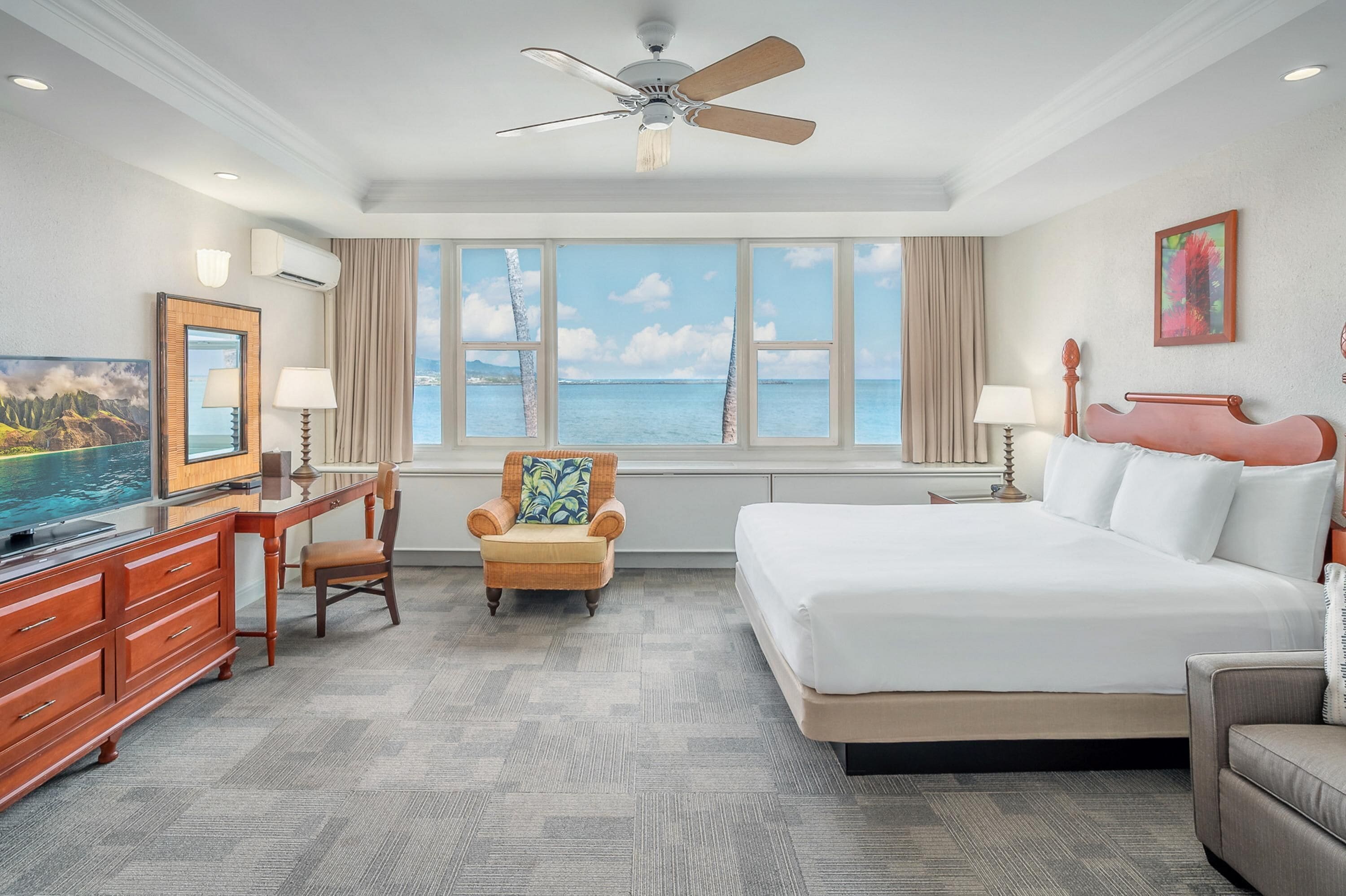 Habitación de hotel con cama, escritorio, silla, televisor y un amplio ventanal con vista al mar.