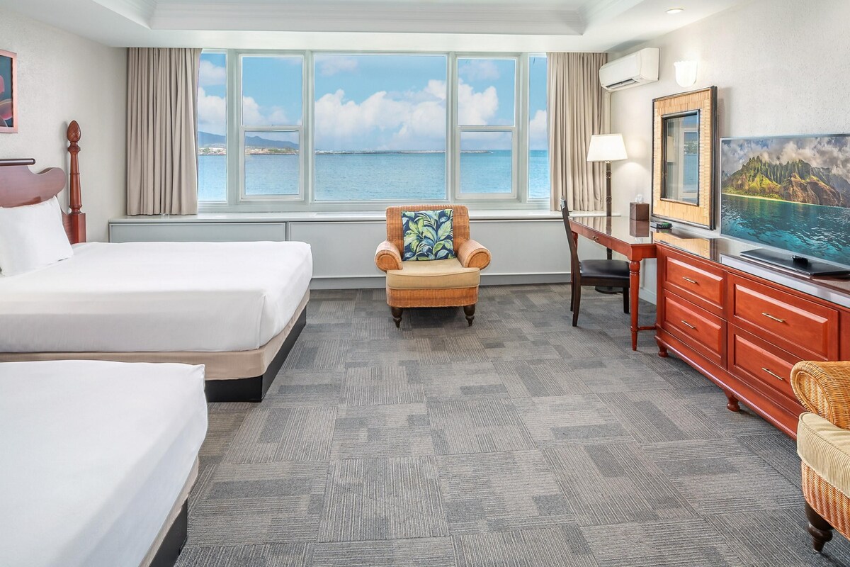 A hotel room with two beds, a chair, a TV, and a large window with a view of the sea.