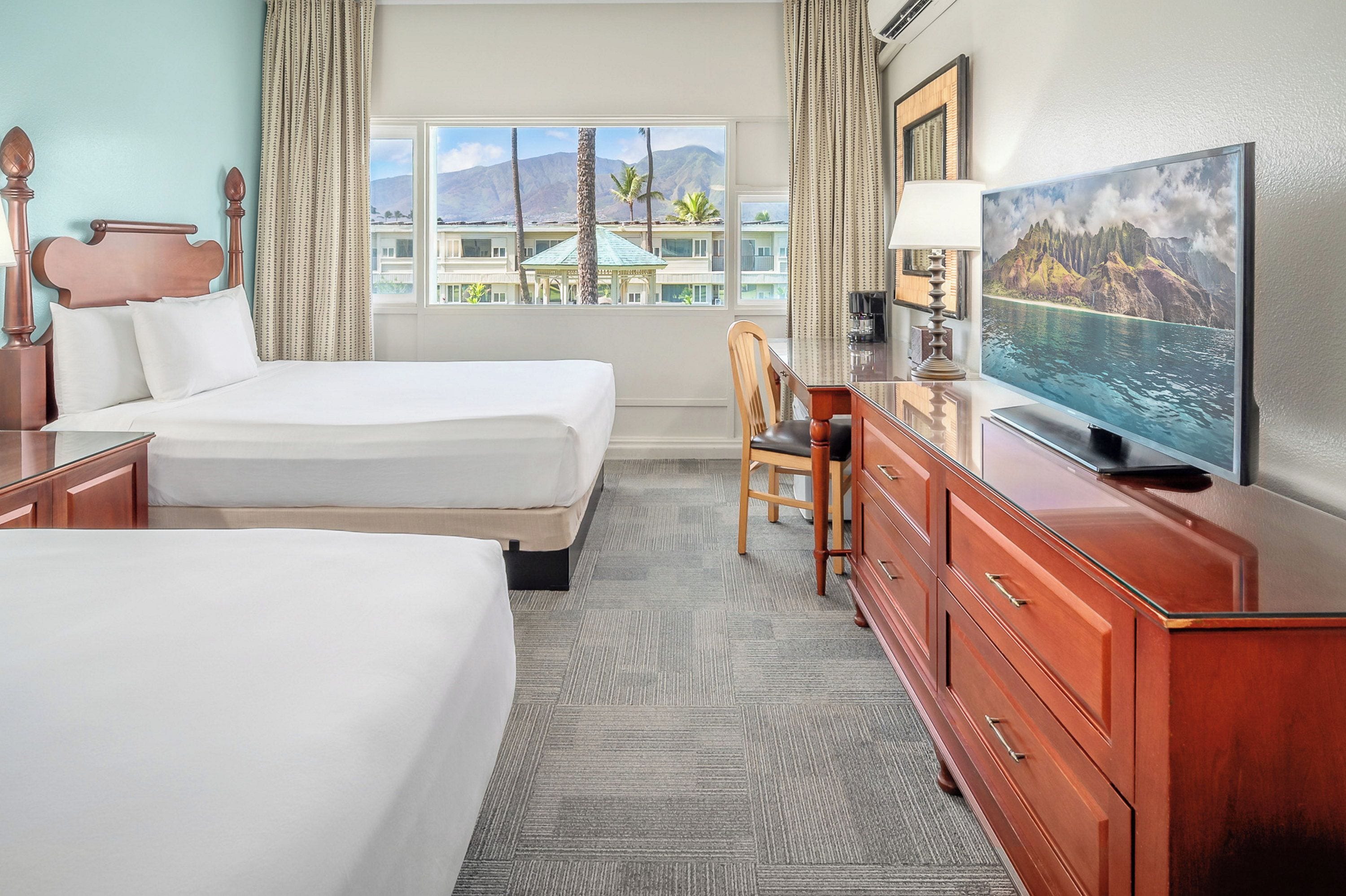 Habitación de hotel con dos camas, televisión, escritorio y vistas a montañas y palmeras.