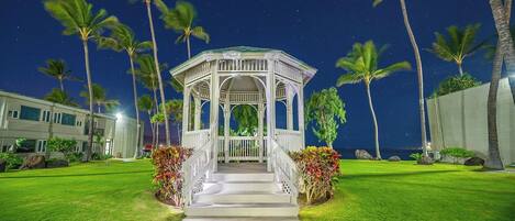 A gazebo with a white exterior and a covered entrance, surrounded by a well-maintained lawn and palm trees.