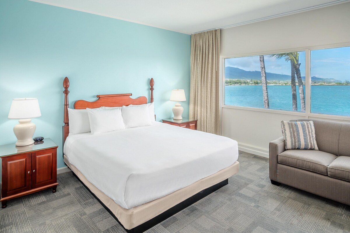A hotel room with a large bed, two bedside tables, a sofa, a window with a view of the sea and palm trees, and a light blue wall.