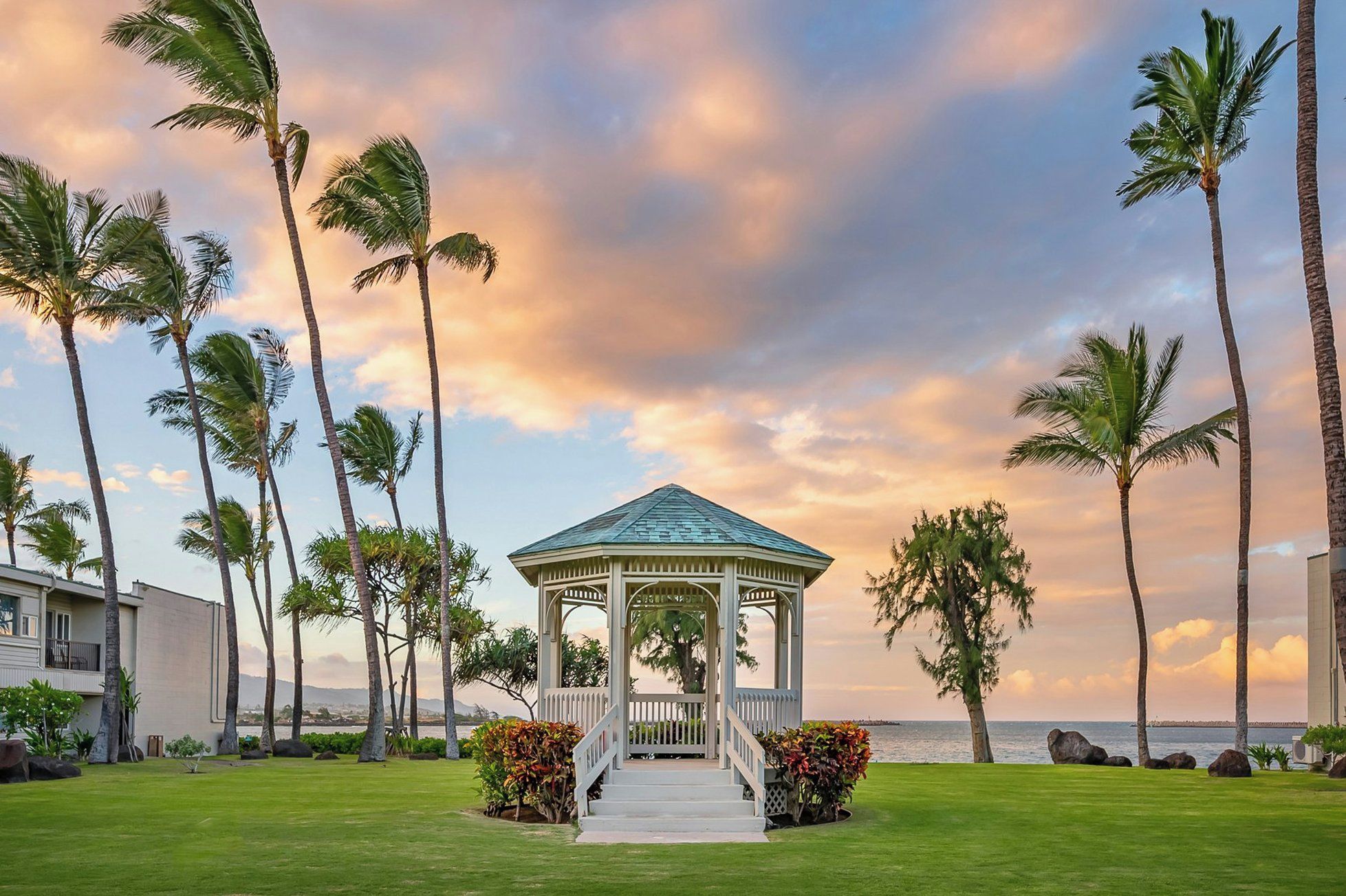 A gazebo with a white roof and open sides, surrounded by a well-maintained lawn and palm trees, with a view of the ocean in the background.