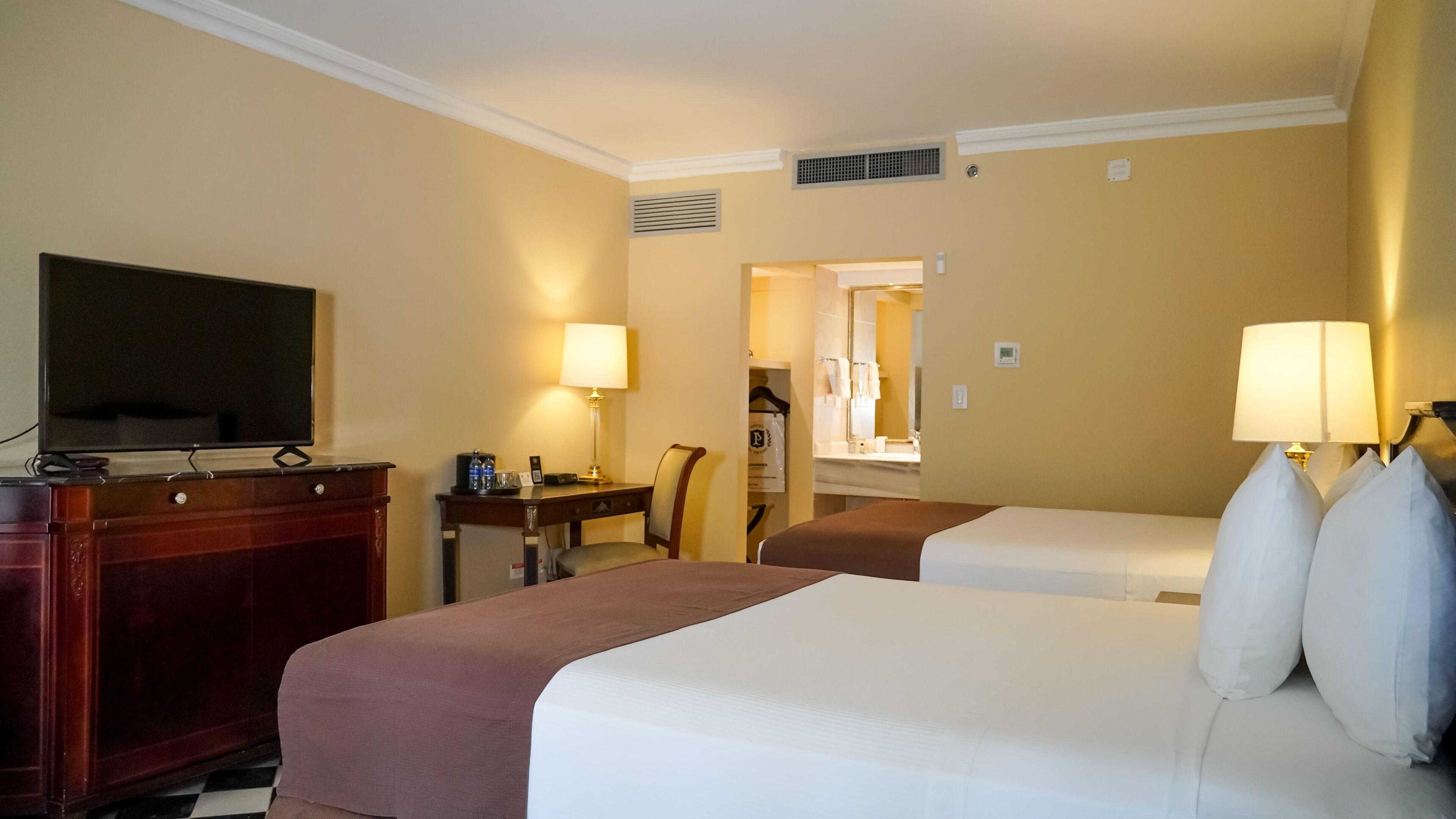 A hotel room with two beds, a desk, a TV, and a bathroom visible through an open door.