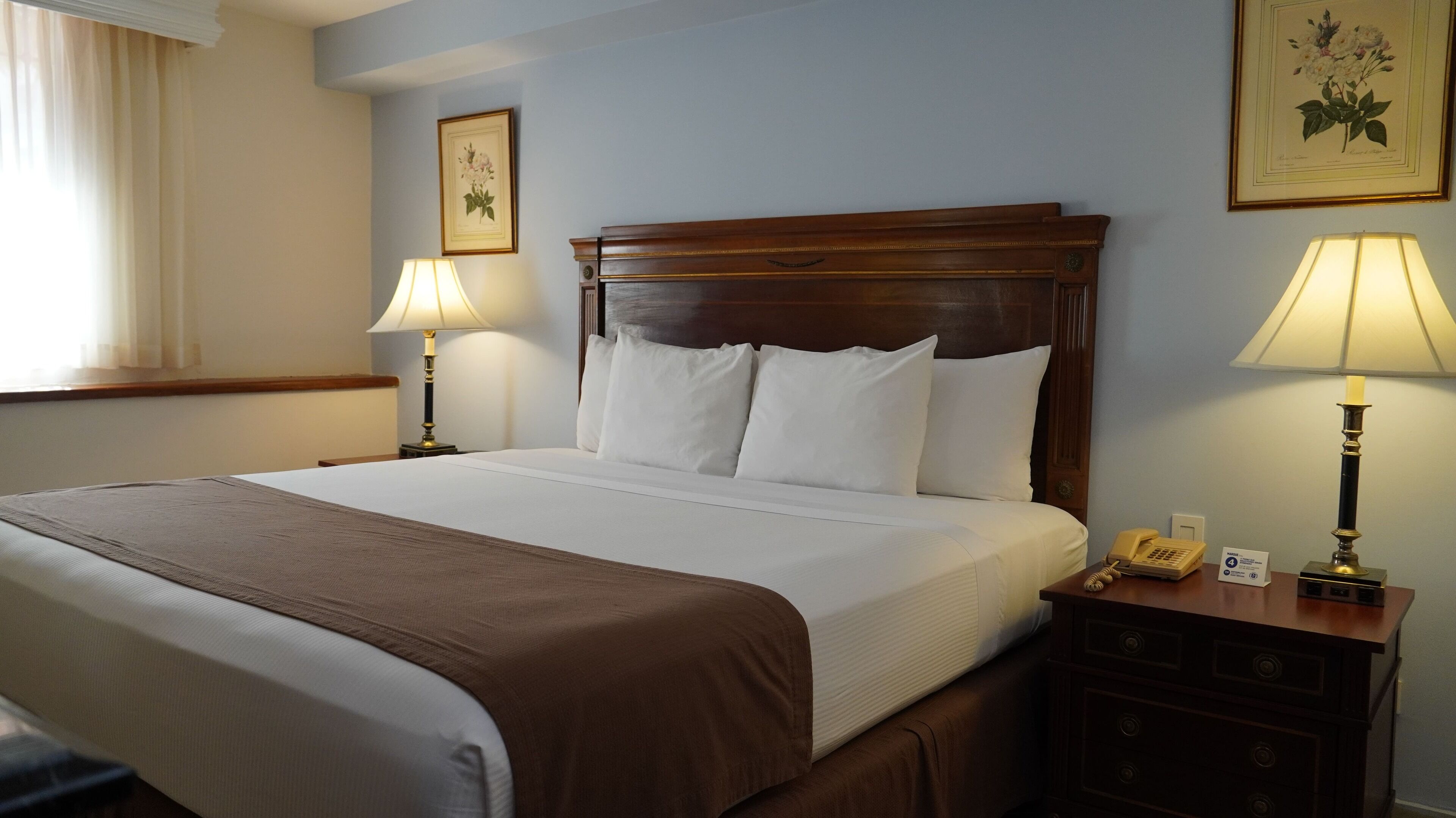 Una habitación de hotel con una cama grande, dos lámparas de noche, una mesita de noche de madera con un teléfono, e impresiones botánicas enmarcadas en las paredes.
