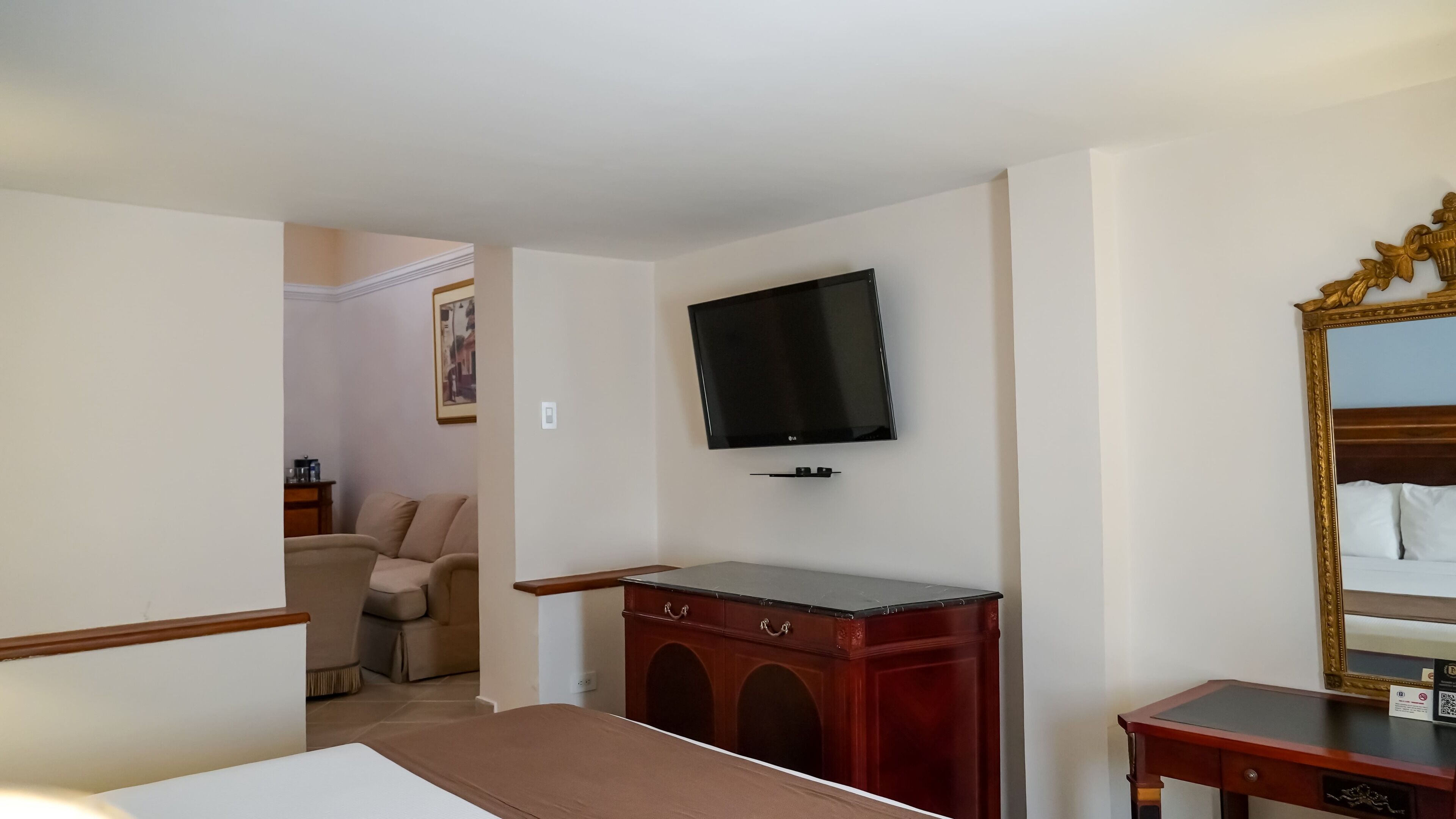 A hotel room with a bed, a TV mounted on the wall, a wooden dresser, and a mirror with a decorative frame.