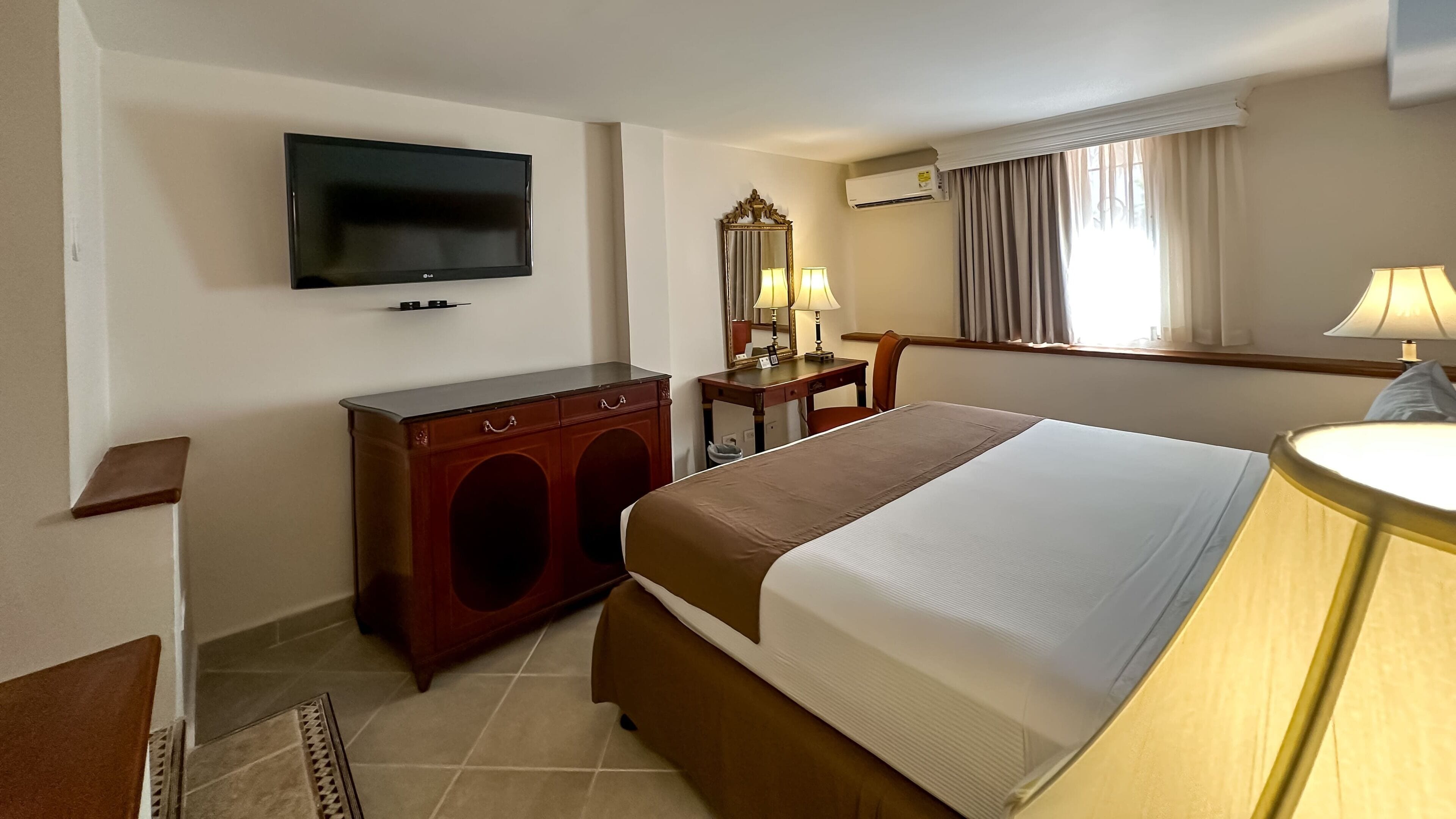 A hotel room with a bed, a TV, a desk with a lamp, and a window with curtains.
