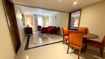 A hotel room with a dining area, a sofa, and a red armchair. There is a small table with a lamp and a mirror on the wall.