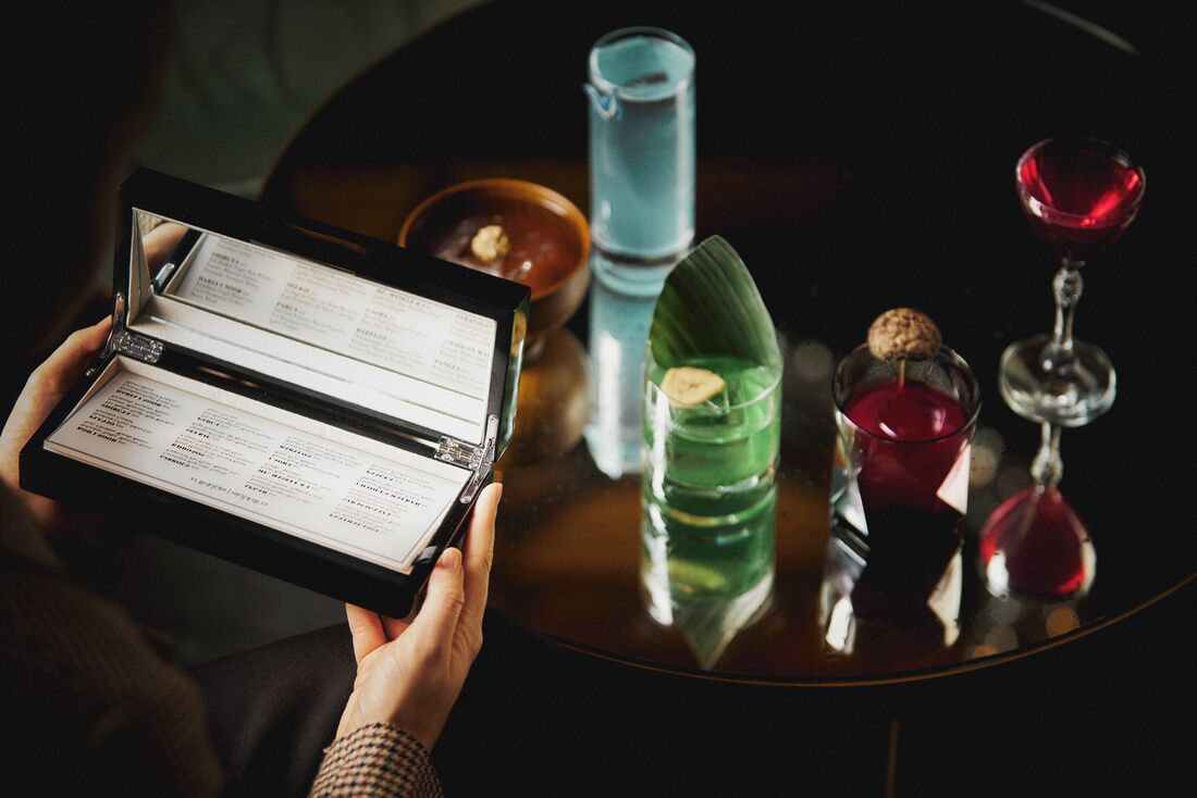 A person holding a menu with a table setting that includes a glass of red wine, a green drink, and a small dish with a nut.