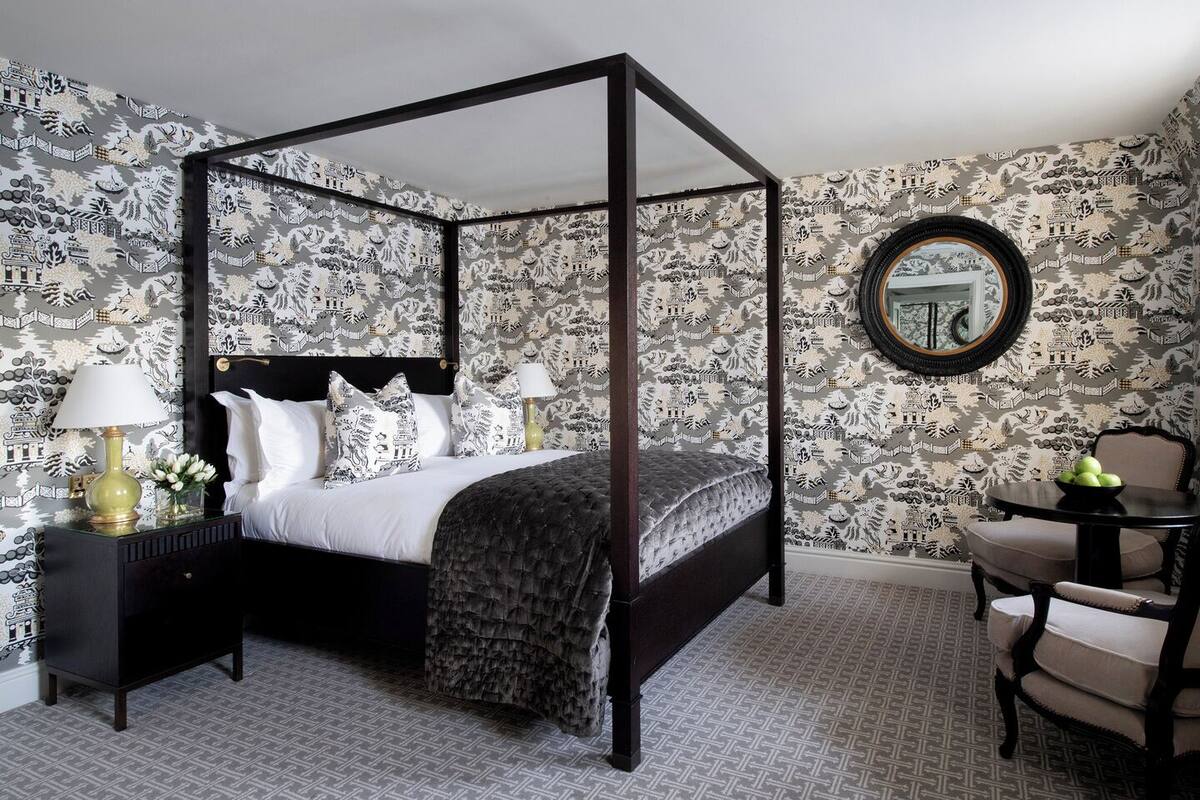 A four-poster bed with a black frame, white bedding, and a patterned throw. There is a round mirror on the wall, a small table with a bowl of fruit, and a chair with a cushion.