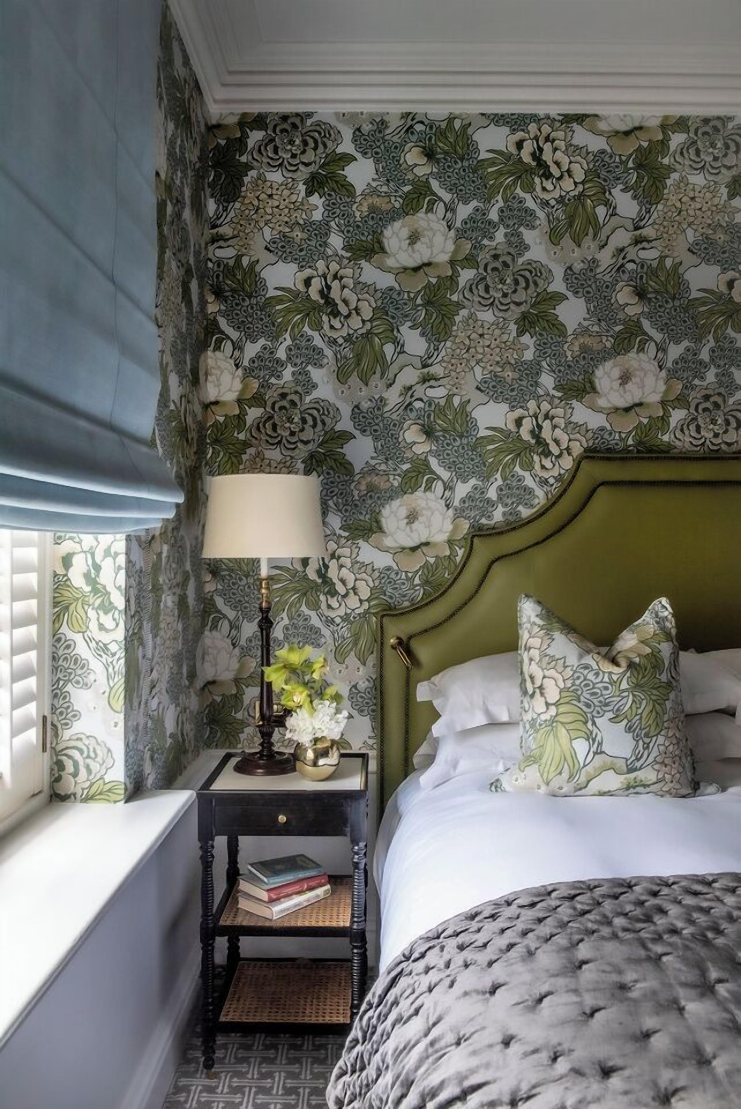 A bedroom with floral wallpaper, a green headboard, a bedside table with a lamp, and a window with blinds.