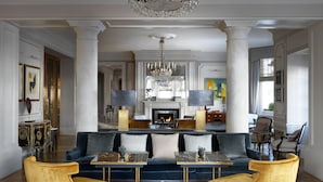 A spacious living room with a blue sofa, gold armchairs, a marble coffee table, and a large chandelier. - The Kensington Hotel (London)