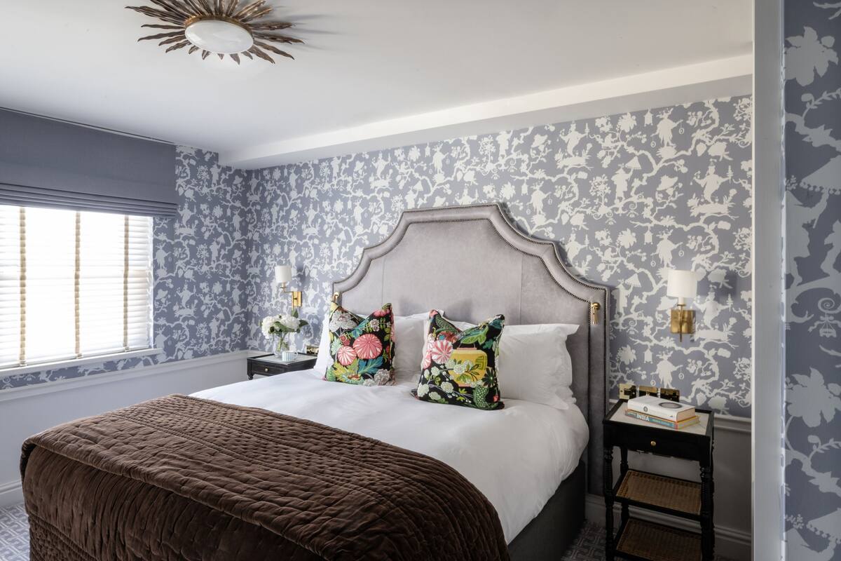 Ein Schlafzimmer mit einem großen Bett, blumenmusterigem Wandpapier, einer Zierleuchte an der Decke und einem Fenster mit Jalousien.