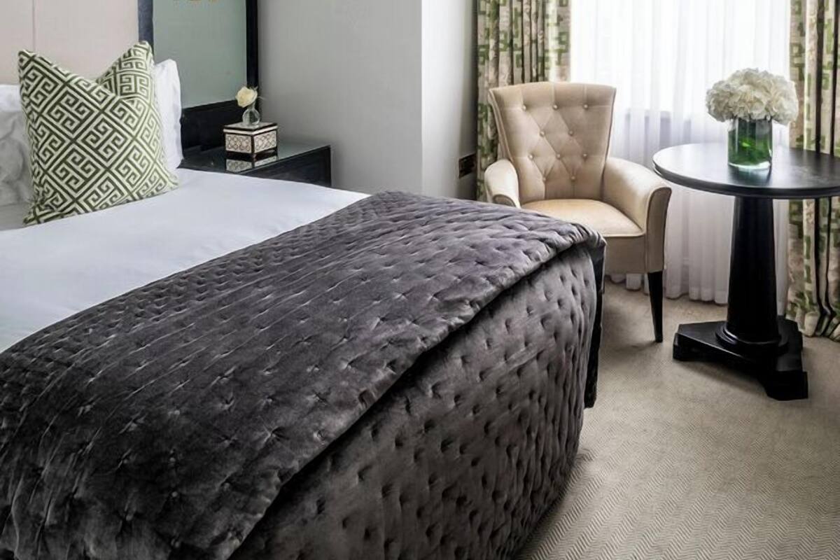 A hotel room with a large bed, a chair, a small table with a vase, and a window with curtains.