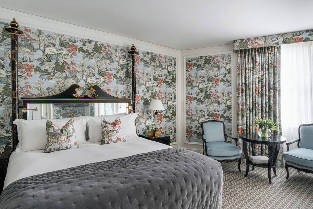 A hotel room with a large bed, a nightstand, a chair, a small table, and a window with curtains.