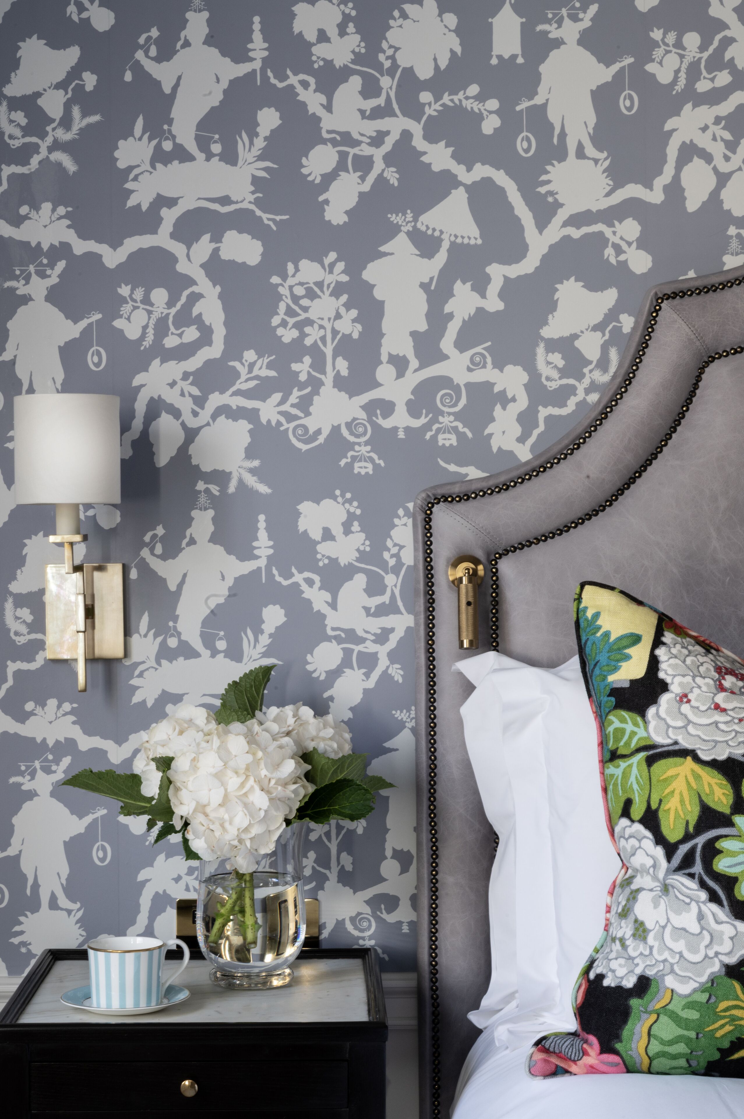 A bedroom with a grey floral wallpaper, a bedside table with a lamp, a vase with flowers, and a striped cup.