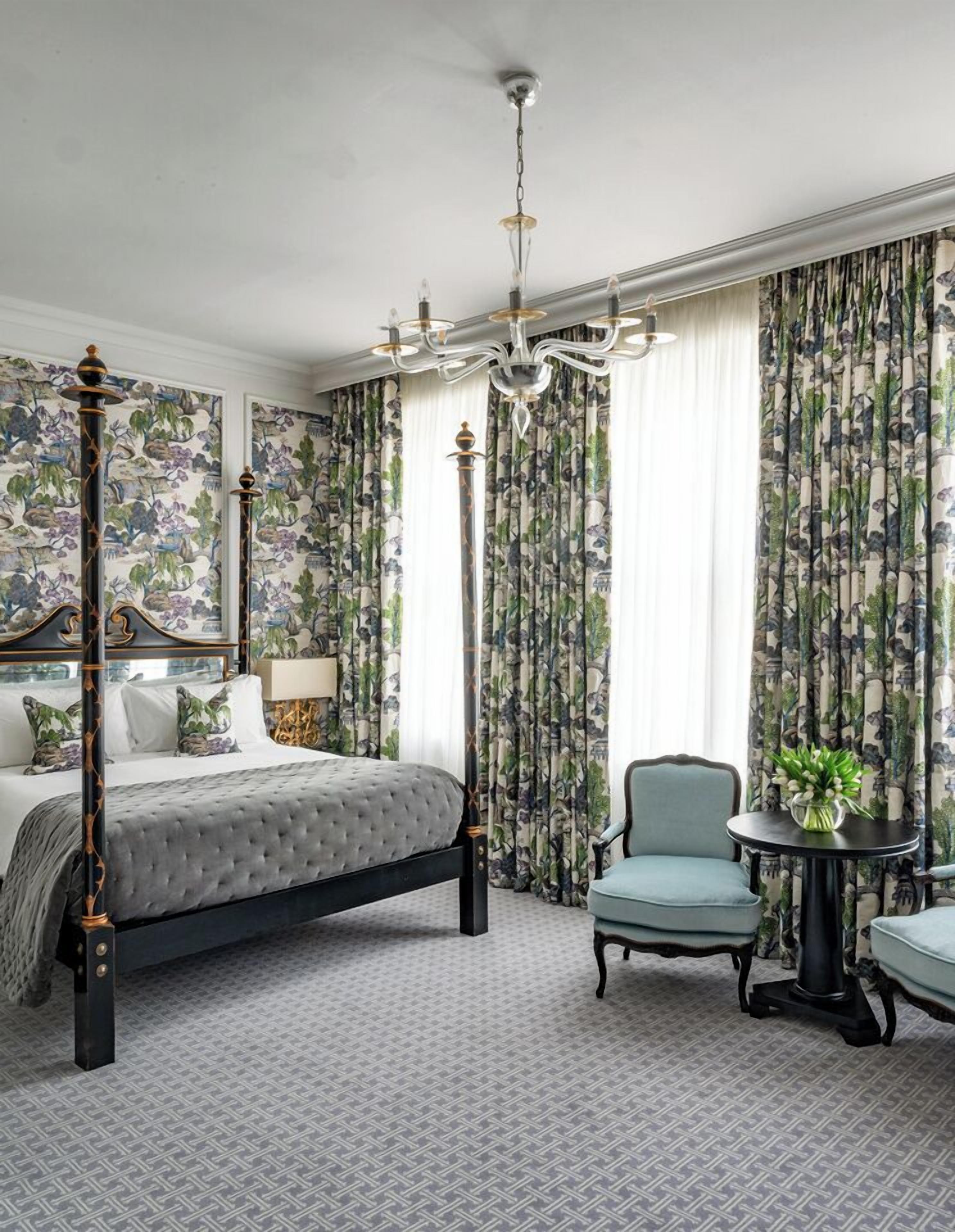 A bedroom with a four-poster bed, floral wallpaper, a chandelier, and a small round table with a plant.