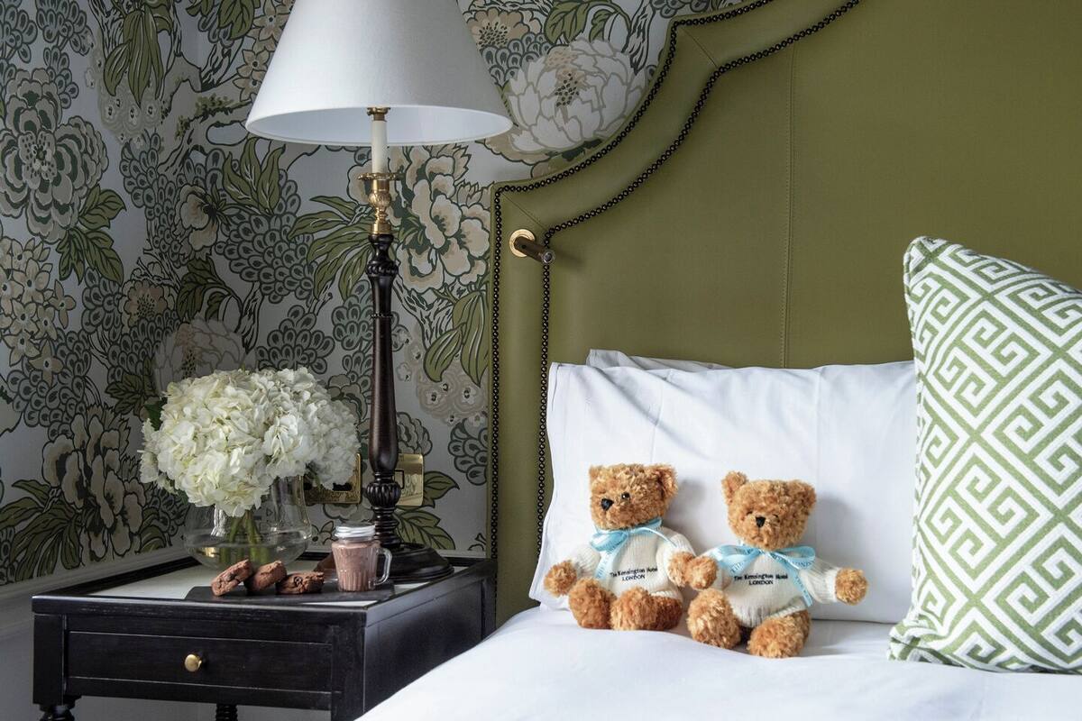A neatly made bed with two teddy bears, a nightstand with a lamp, a vase of flowers, and a patterned pillow.