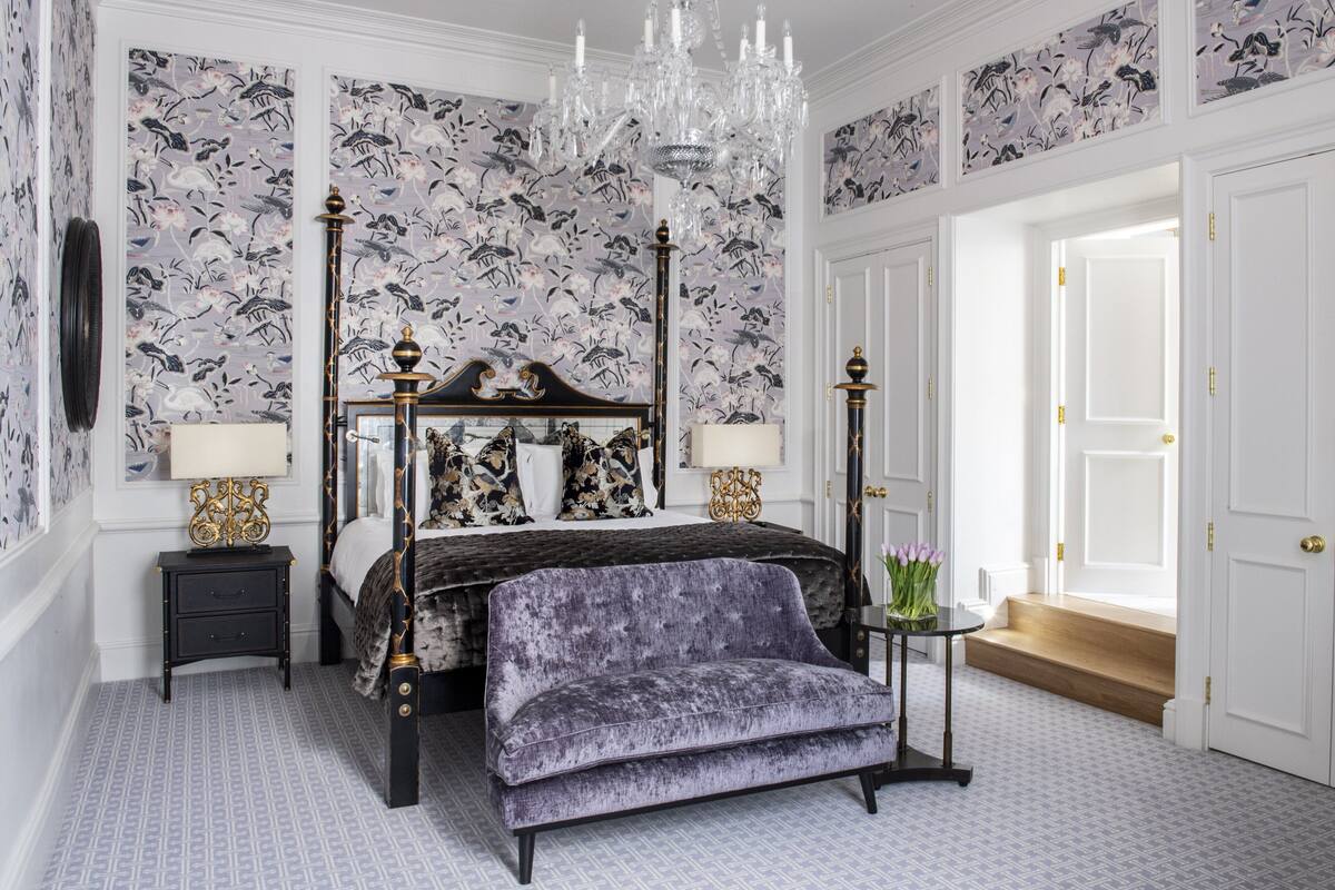 A bedroom with a four-poster bed, a chandelier, floral wallpaper, and a bench.