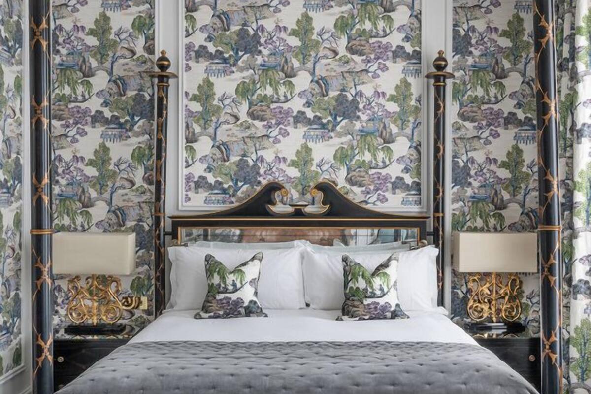 Ein Bett mit Baldachin, dessen dunkler Rahmen goldene Akzente aufweist, wird von zwei Nachttischen mit Lampen begleitet. Der Raum zeichnet sich durch einen Wandteppich mit floralen Mustern und einen strukturierten Boden aus.