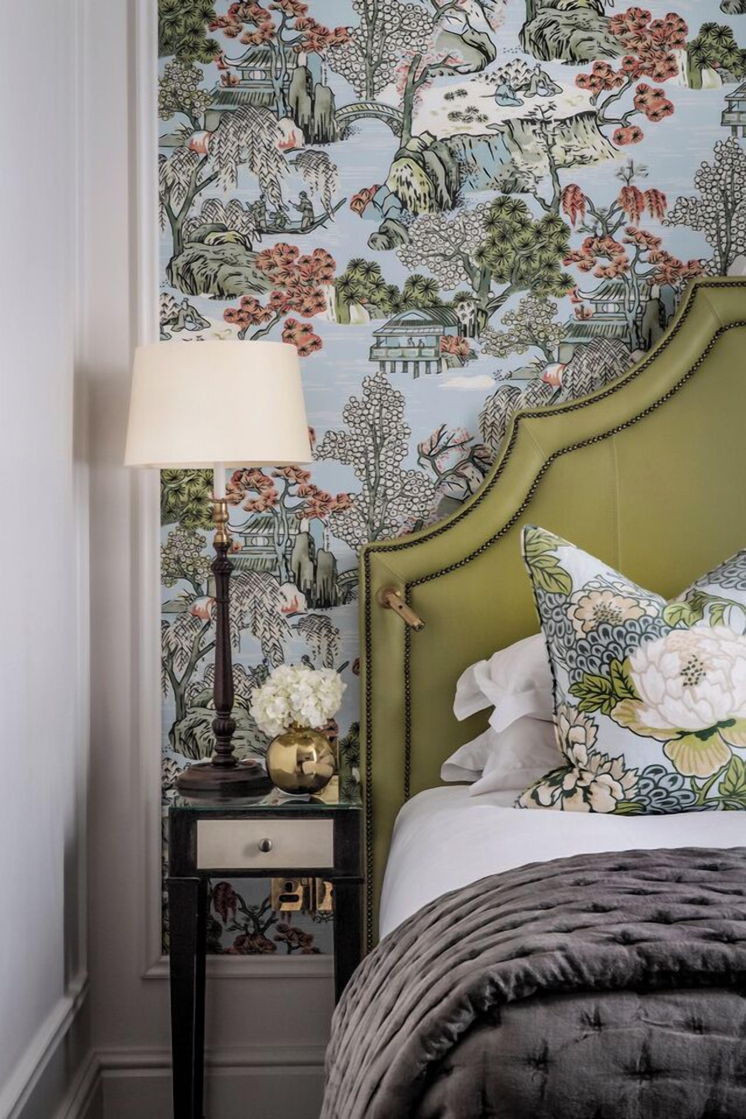 Un dormitorio con cabecera tapizada en verde, una mesita de noche con lámpara y papel tapiz estampado con motivos florales.