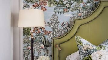 Ein Schlafzimmer mit einem grünen, gepolsterten Kopfteil, einem Nachttisch mit Lampe und blumenmuster-tapetierter Wand.